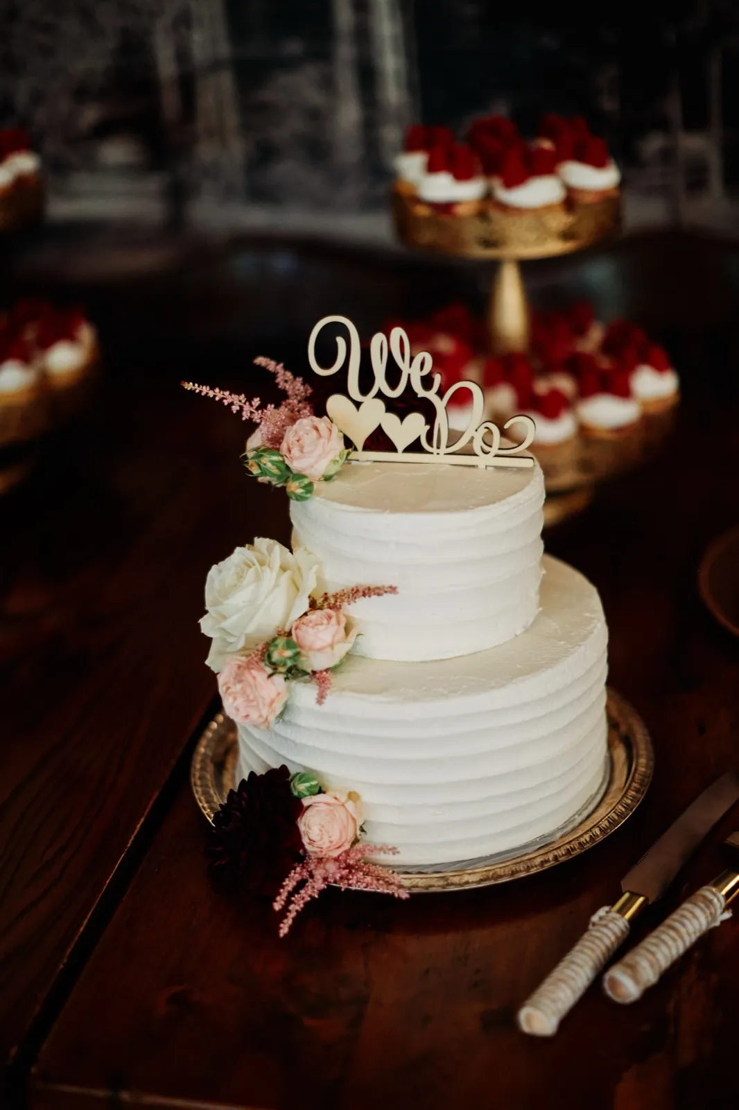 A two-tier white wedding cake with a 'We Do' topper and blush and burgundy florals sits on a silver platter, with cupcakes and cake knives blurred in the background. — Tim Larsen Photography, Brainerd Lakes MN
