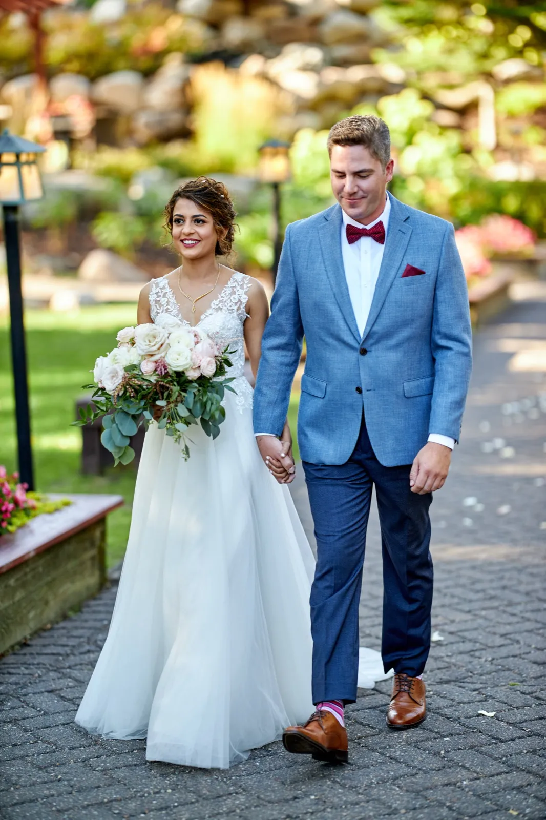 The bride and groom walk hand in hand down a stone garden path at Grand View Lodge, the bride holding a lush blush and white bouquet while the groom looks down with a soft smile. — Tim Larsen Photography, Brainerd Lakes MN