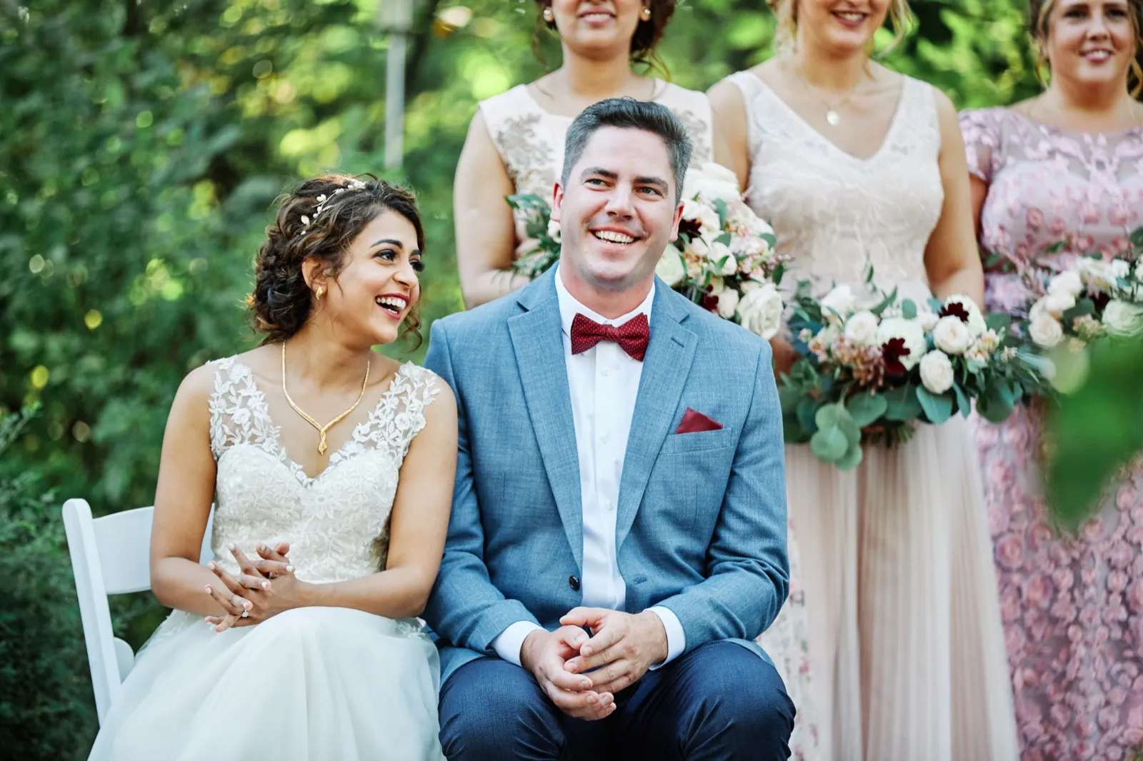 The bride laughs while looking off to the side as the groom grins toward the camera, both seated during their outdoor ceremony with bridesmaids holding bouquets standing behind them. — Tim Larsen Photography, Brainerd Lakes MN