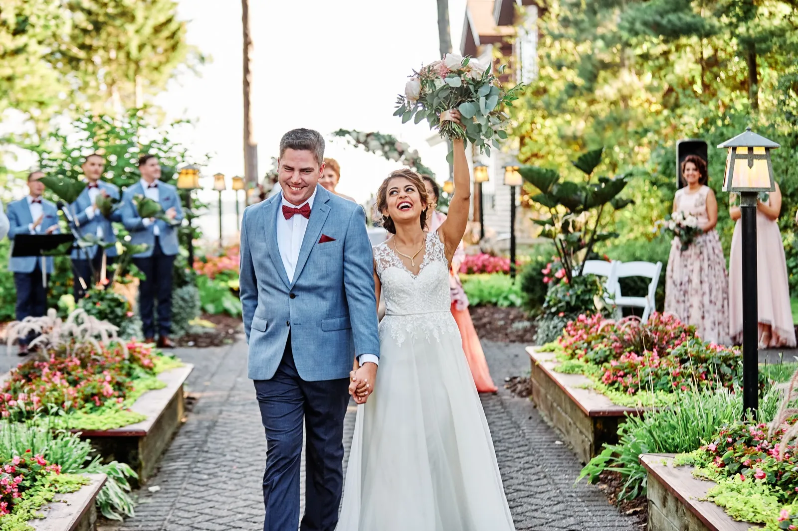 A bride raises her bouquet overhead and laughs while walking hand-in-hand with the groom down a garden aisle, flanked by their bridal party at Grand View Lodge. — Tim Larsen Photography, Brainerd Lakes MN