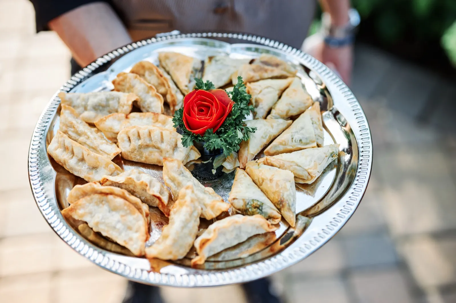 A server holds a silver platter of fried dumplings and samosas arranged around a carved tomato rose garnish. — Tim Larsen Photography, Brainerd Lakes MN
