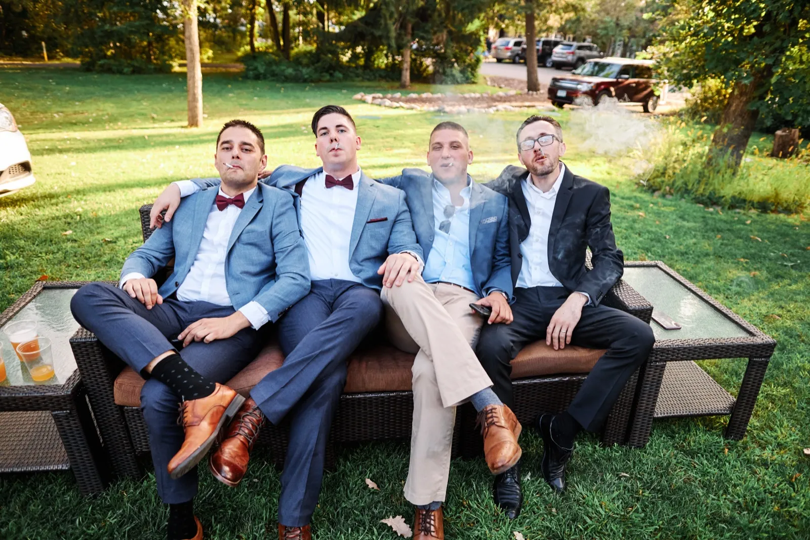 The groom and three groomsmen sit shoulder-to-shoulder on an outdoor wicker couch, smoking cigars and looking deadpan at the camera as smoke drifts around them. — Tim Larsen Photography, Brainerd Lakes MN