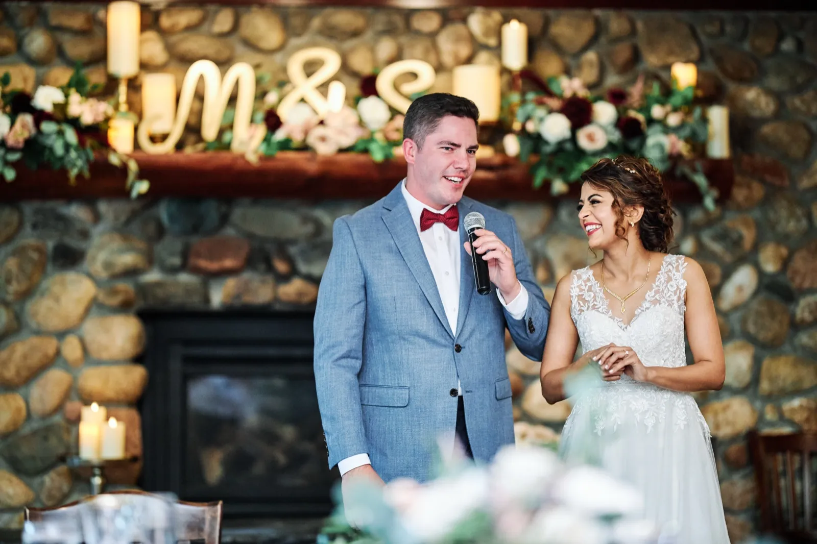 The groom speaks into a microphone while the bride looks up at him laughing, both standing beneath a stone fireplace mantel decorated with florals, candles, and a lit M&E sign. — Tim Larsen Photography, Brainerd Lakes MN