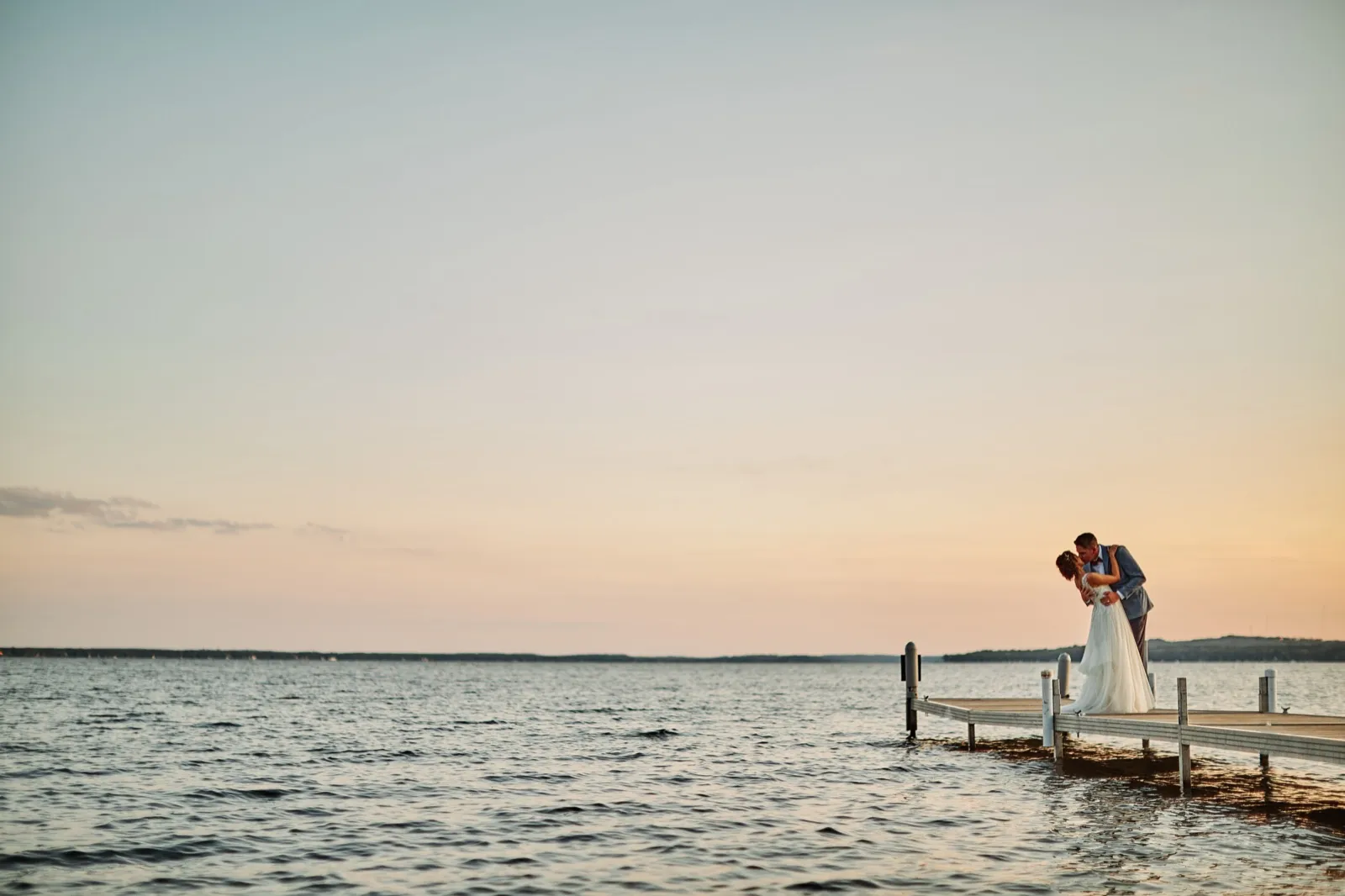 The bride and groom share a dip-kiss at the end of a wooden dock as the sky fades into pastel dusk over Gull Lake. — Tim Larsen Photography, Brainerd Lakes MN