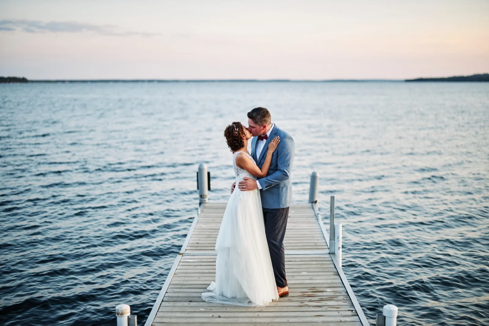 The bride and groom kiss at the end of a long wooden dock extending into Gull Lake under a soft dusk sky. — Tim Larsen Photography, Brainerd Lakes MN