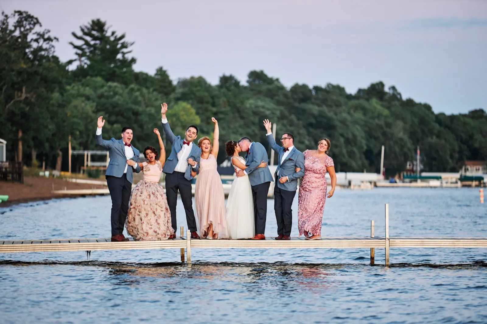The bridal party cheers with arms raised on a dock over Gull Lake while the bride and groom kiss at center during dusk. — Tim Larsen Photography, Brainerd Lakes MN