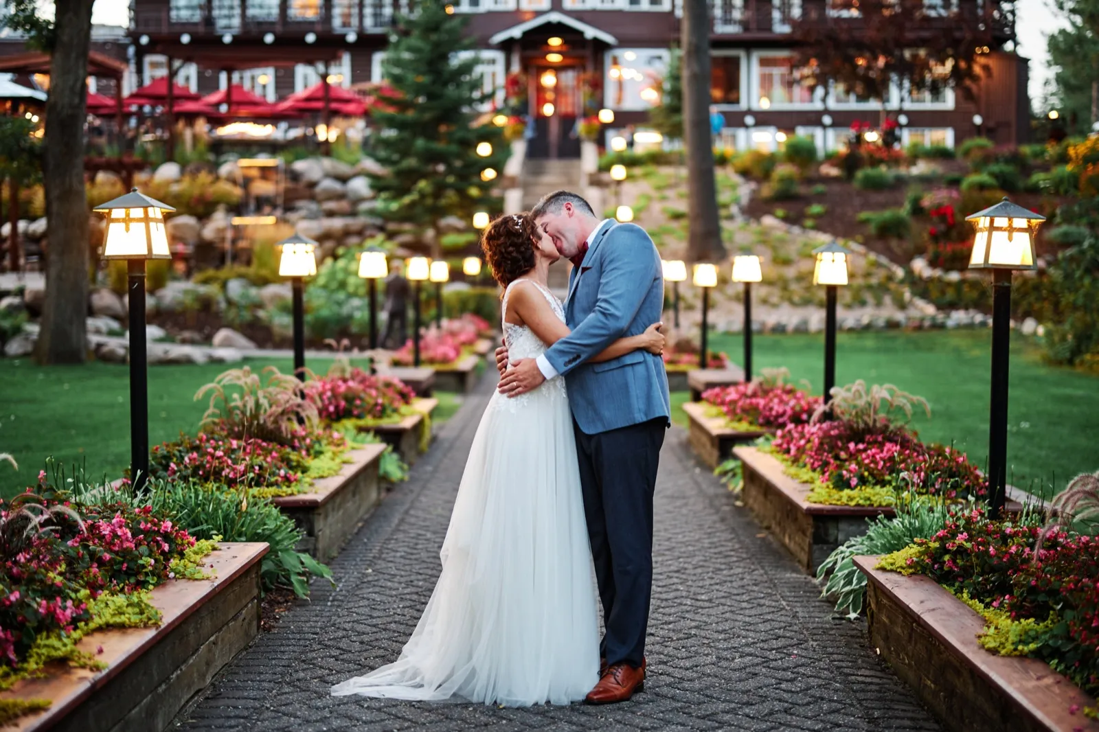 The couple kisses at the center of a symmetrical lantern-lined garden path at dusk, with the illuminated Grand View Lodge rising behind them. — Tim Larsen Photography, Brainerd Lakes MN