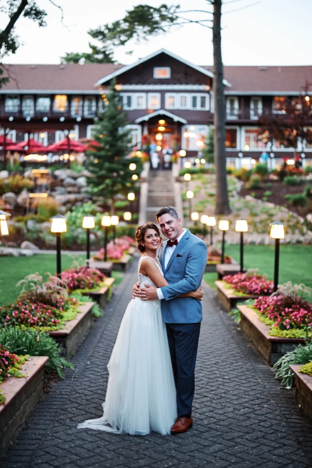 The couple embraces on a stone path lined with glowing lanterns and flower beds, with the illuminated Grand View Lodge facade rising behind them at dusk. — Tim Larsen Photography, Brainerd Lakes MN
