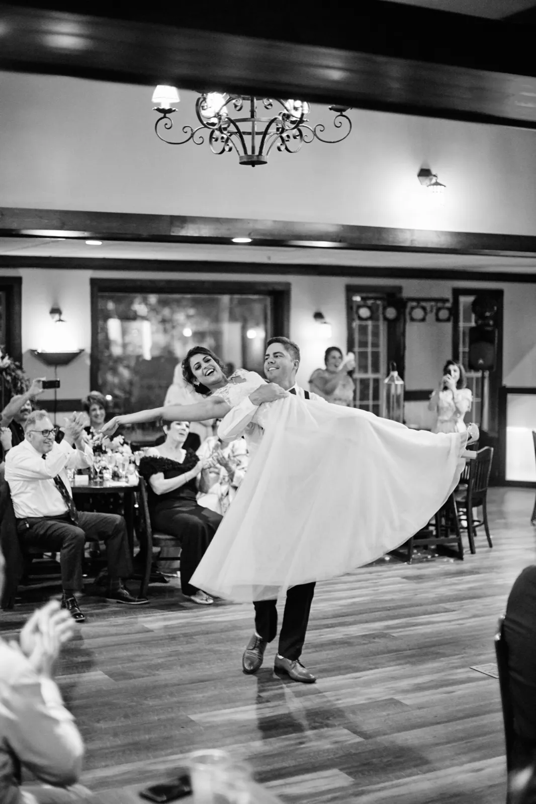 The groom spins the bride mid-air on the dance floor, her dress flaring wide as guests applaud from surrounding tables under a wrought-iron chandelier. — Tim Larsen Photography, Brainerd Lakes MN