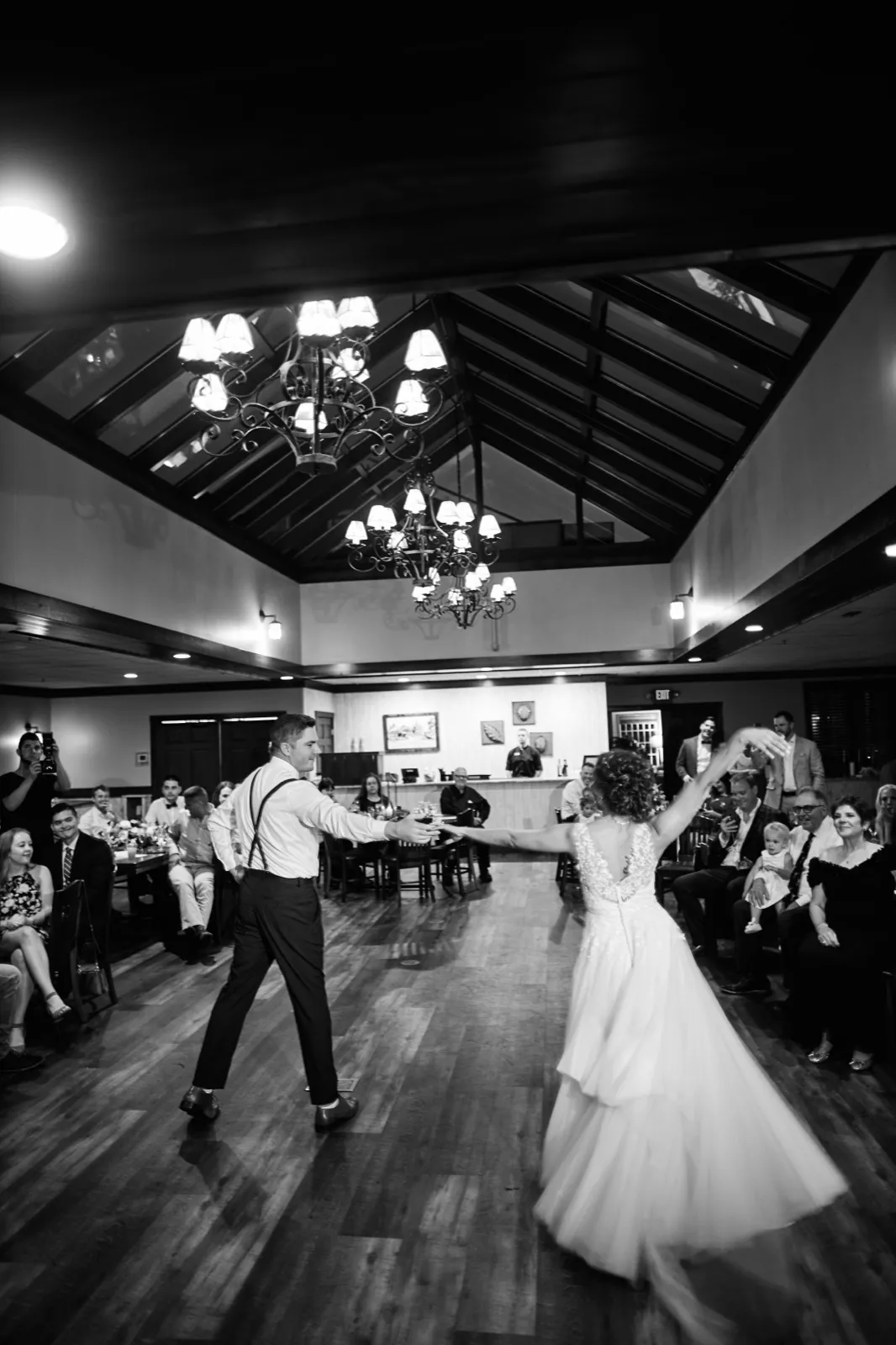 The bride and groom dance together on a wooden floor beneath vaulted ceilings and ornate chandeliers as seated guests watch from both sides of the room. — Tim Larsen Photography, Brainerd Lakes MN