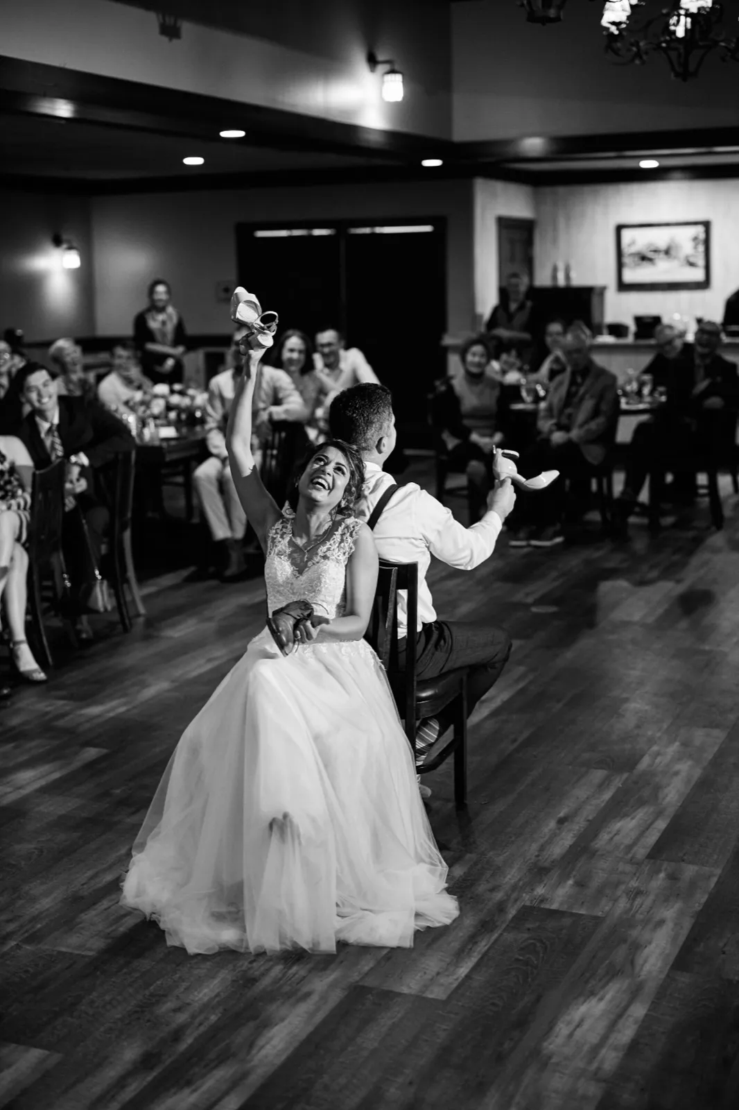 The bride laughs and raises a shoe overhead while seated back-to-back with the groom during a shoe game in front of seated wedding guests. — Tim Larsen Photography, Brainerd Lakes MN