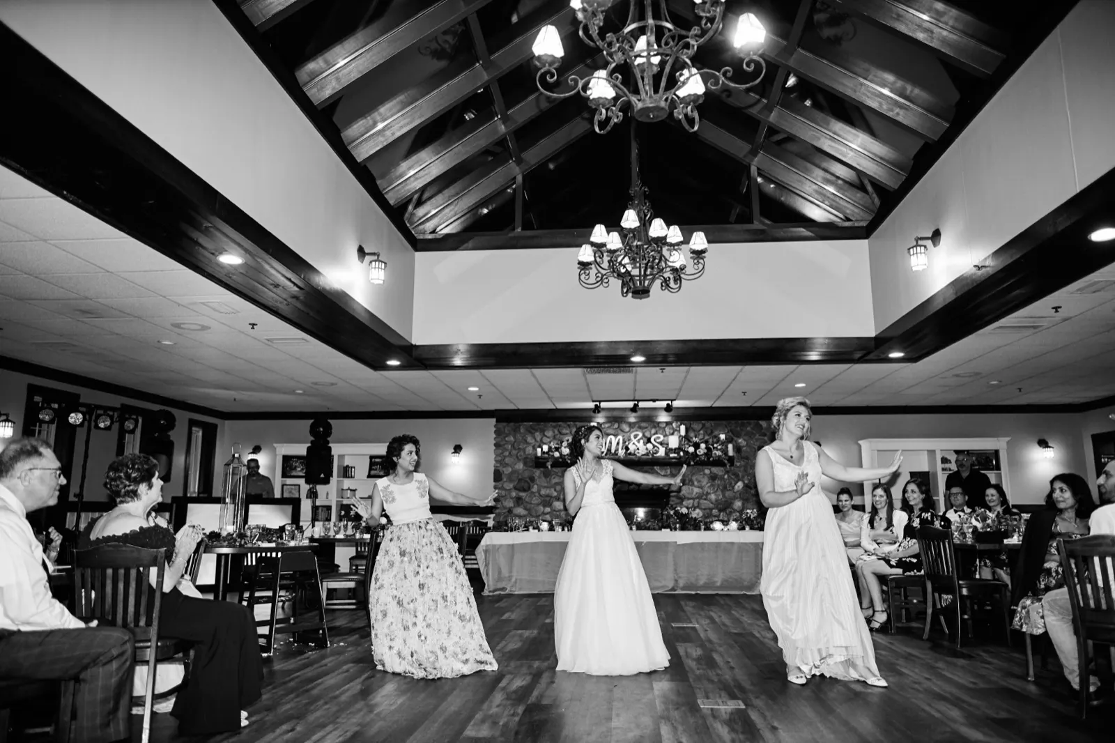 A wide black-and-white view of the bride and two bridesmaids dancing on the floor of a vaulted reception hall while seated guests watch from the perimeter. — Tim Larsen Photography, Brainerd Lakes MN