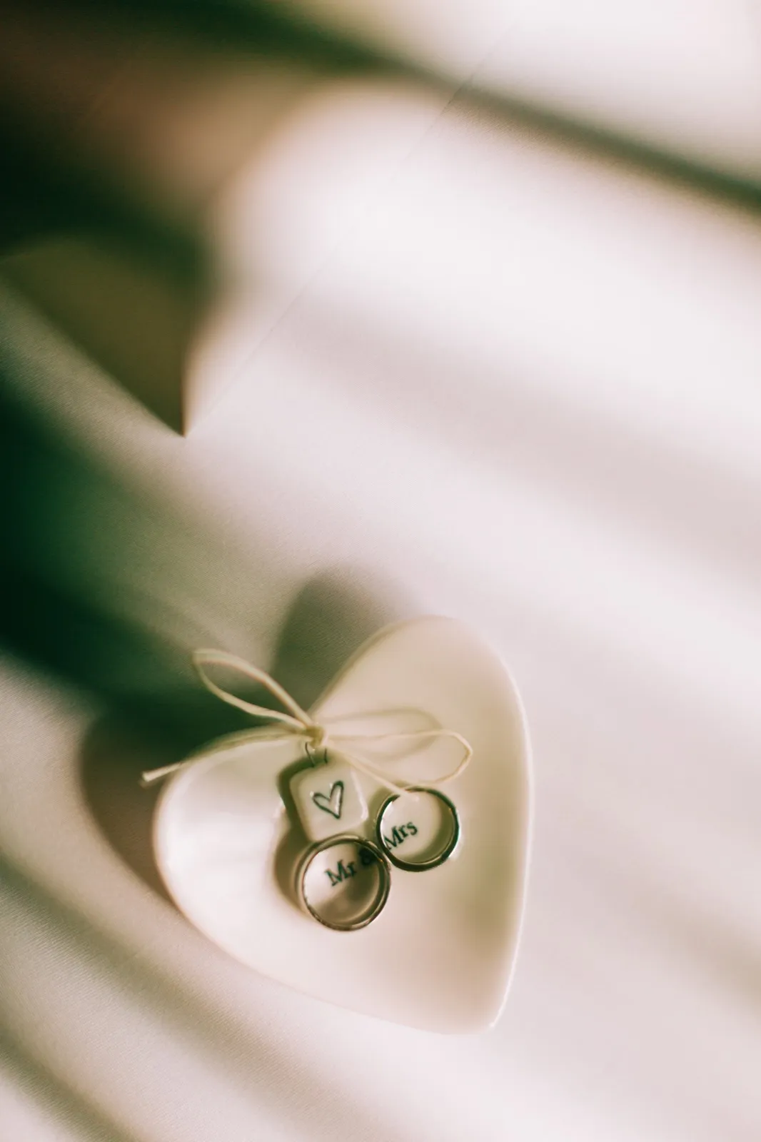 Katie and Brenden's wedding rings rest in a small heart-shaped dish on folded ivory fabric in window light — Tim Larsen Photography, Brainerd Lakes MN