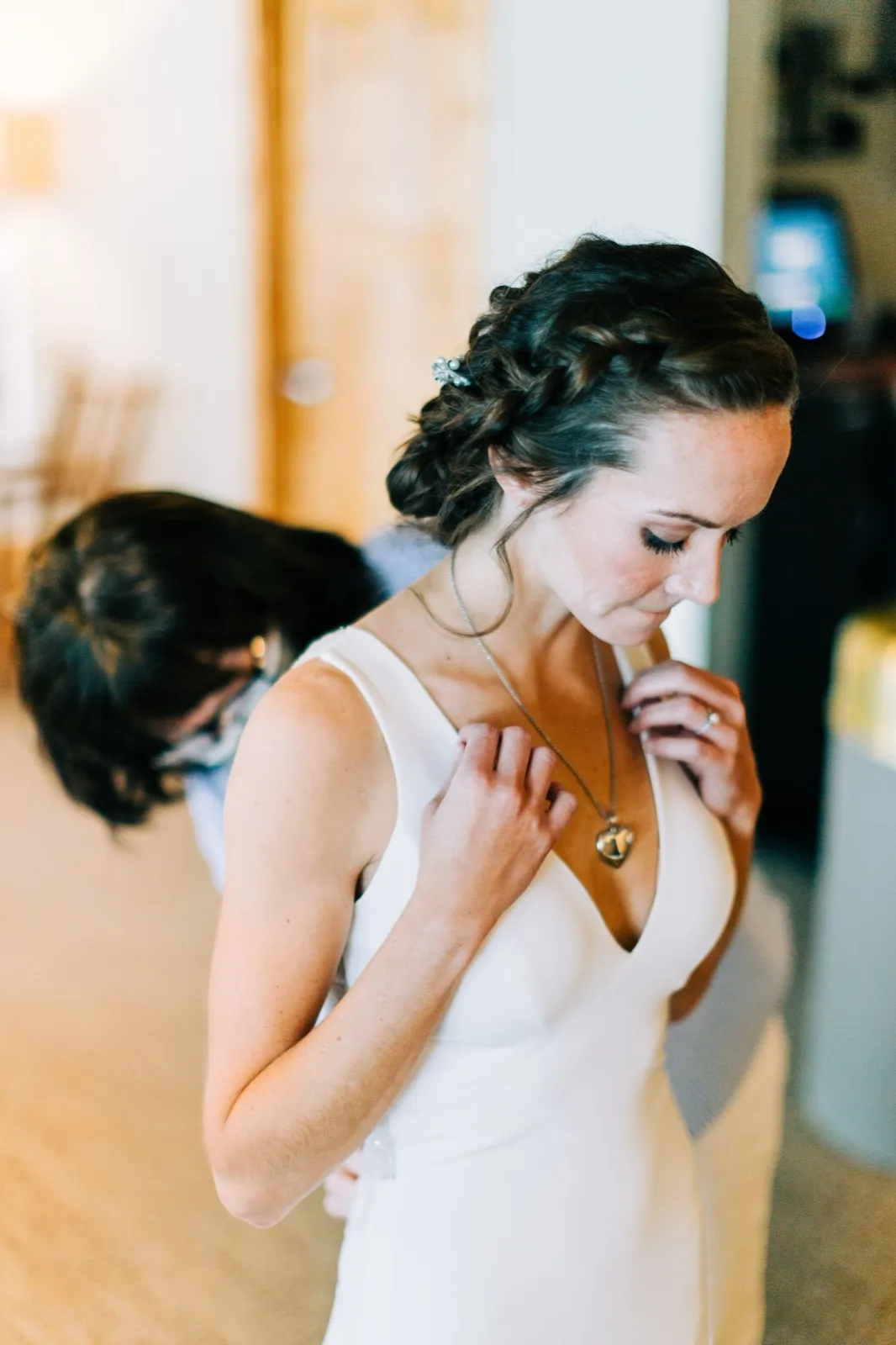 Katie holds a heart-shaped pendant to her chest, eyes downcast, as an attendant zips her gown in the window-lit bridal suite — Tim Larsen Photography, Brainerd Lakes MN