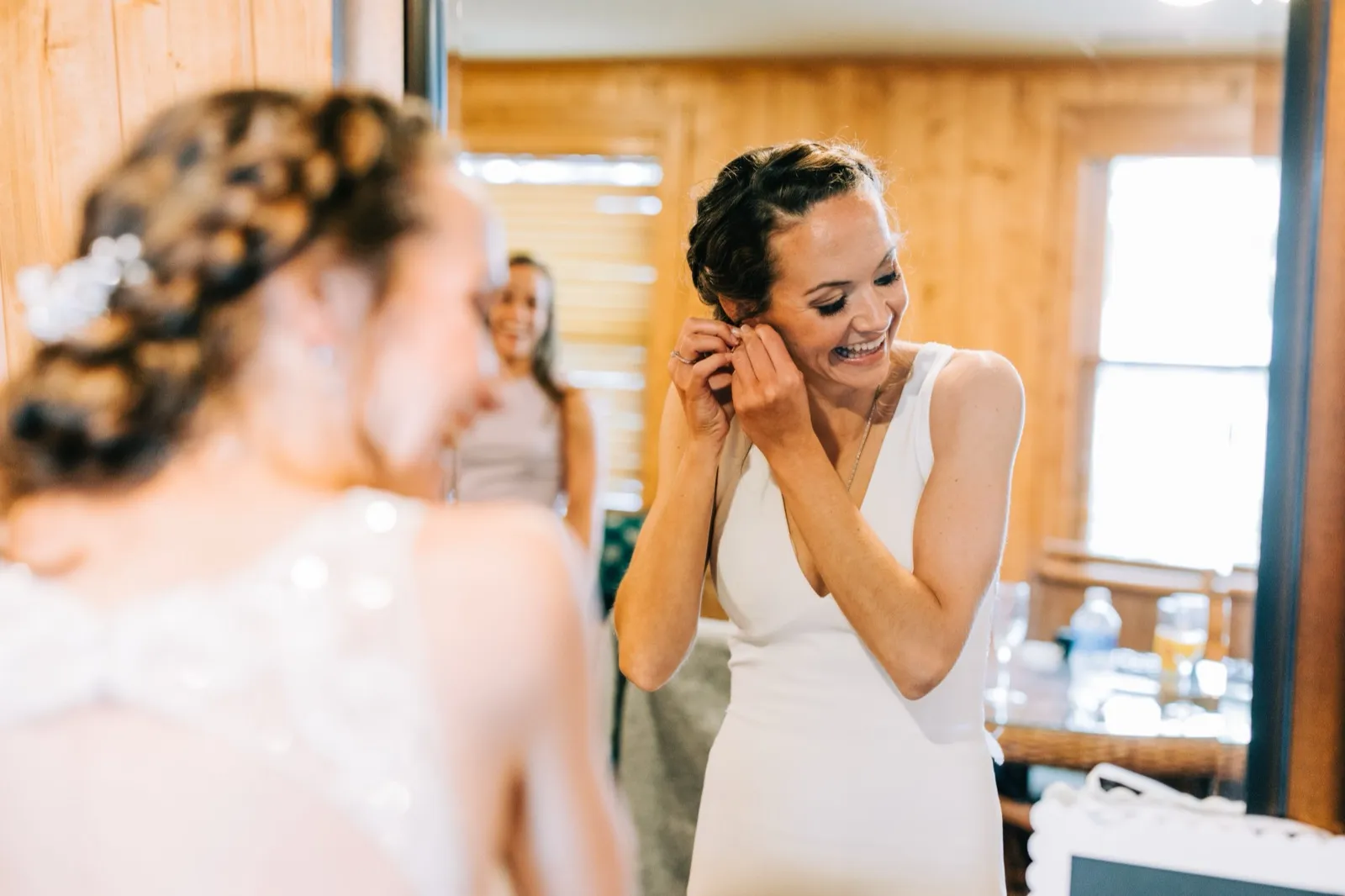Katie laughs while fastening an earring at the mirror, a bridesmaid reflected behind her — Tim Larsen Photography, Brainerd Lakes MN