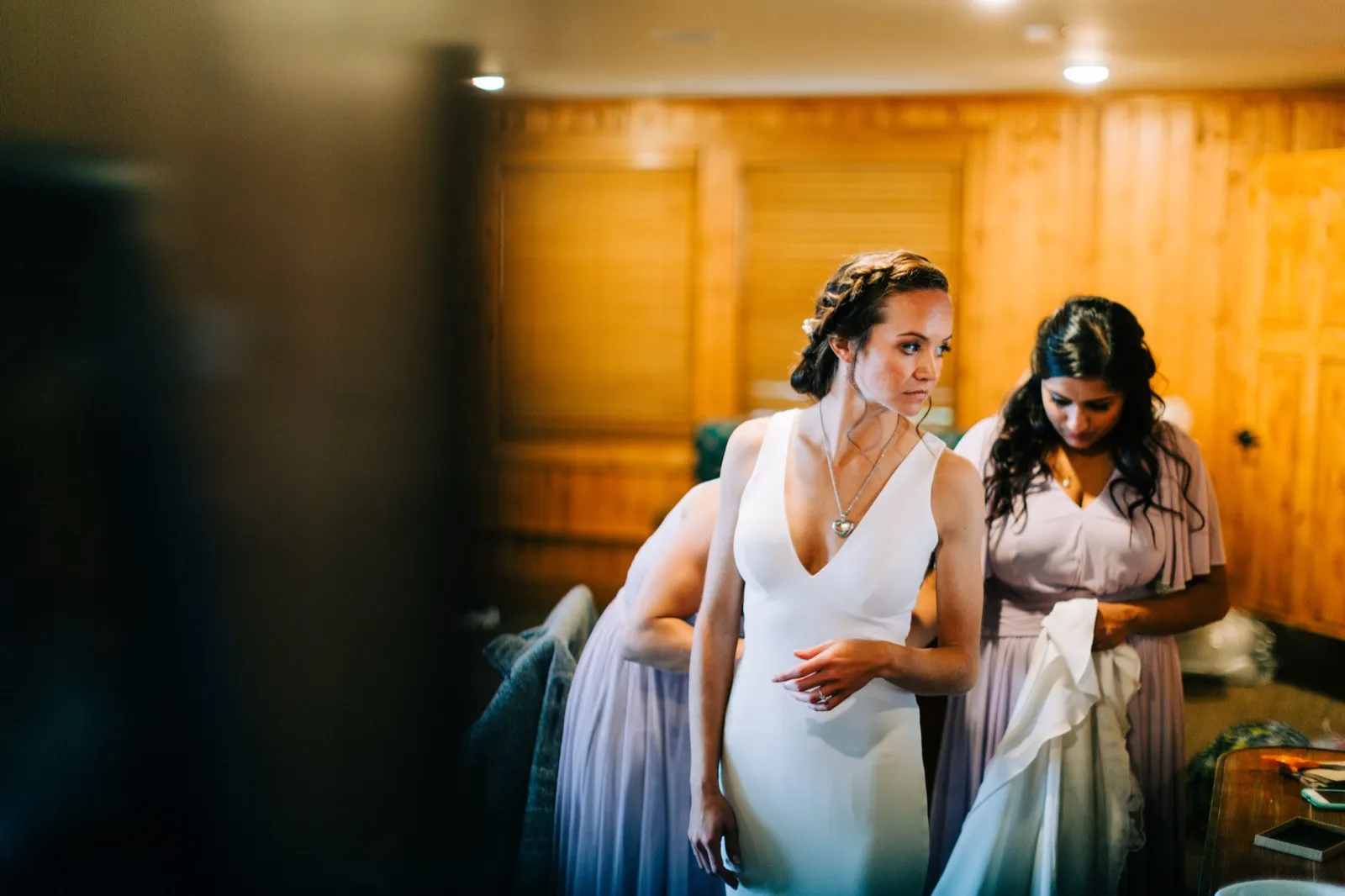 Two bridesmaids in lavender help with Katie's gown in a wood-paneled getting-ready room at Madden's — Tim Larsen Photography, Brainerd Lakes MN