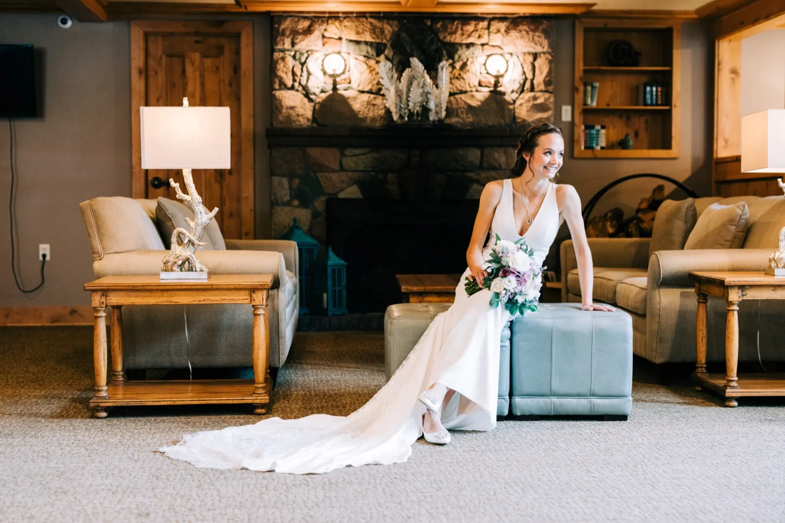 Katie sits on a blue tufted ottoman before a stone fireplace, her train fanned across the carpet as she smiles at someone out of frame — Tim Larsen Photography, Brainerd Lakes MN