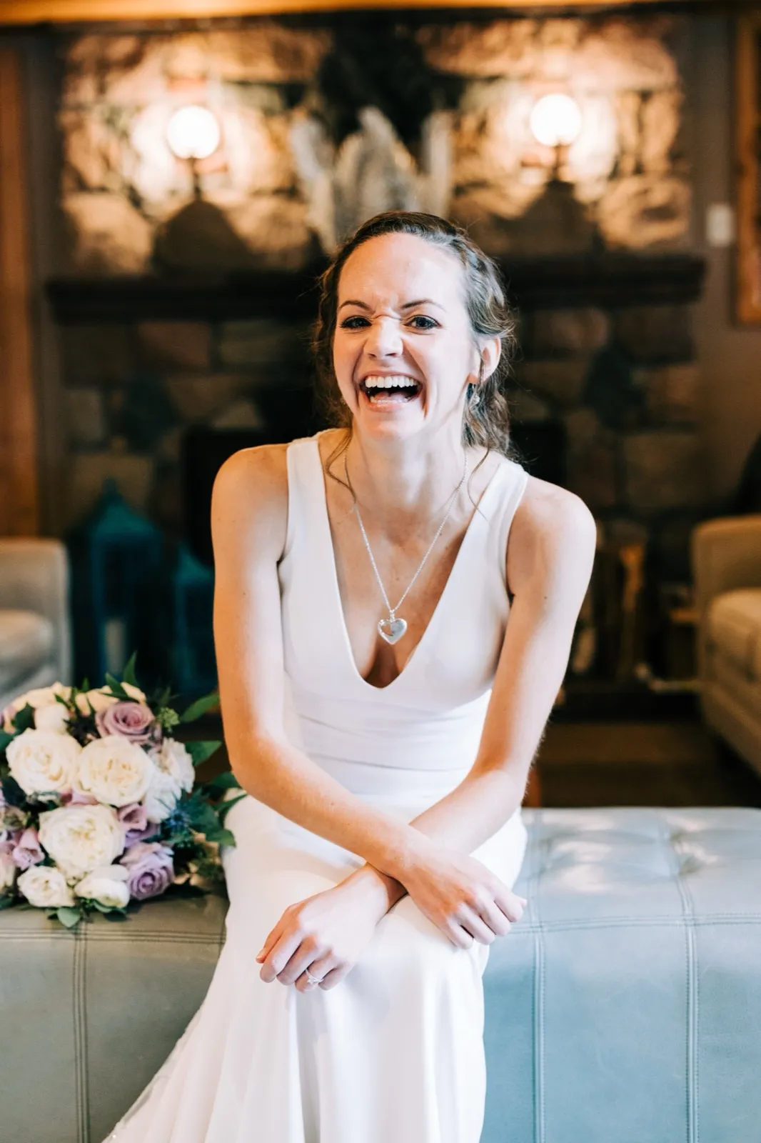 Katie laughs openly on a leather ottoman before a stone fireplace, her cream-and-lavender bouquet beside her — Tim Larsen Photography, Brainerd Lakes MN