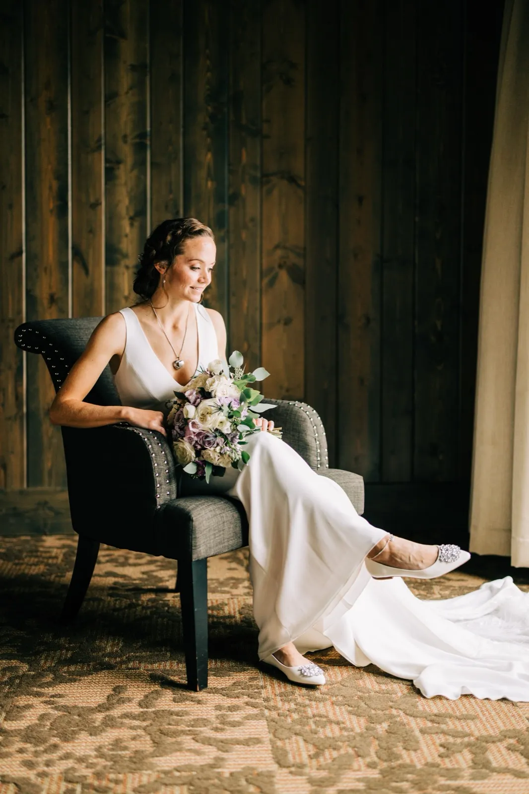 Katie sits in a gray armchair with her lavender-and-cream bouquet, gazing toward the window, train spilling across the rug — Tim Larsen Photography, Brainerd Lakes MN