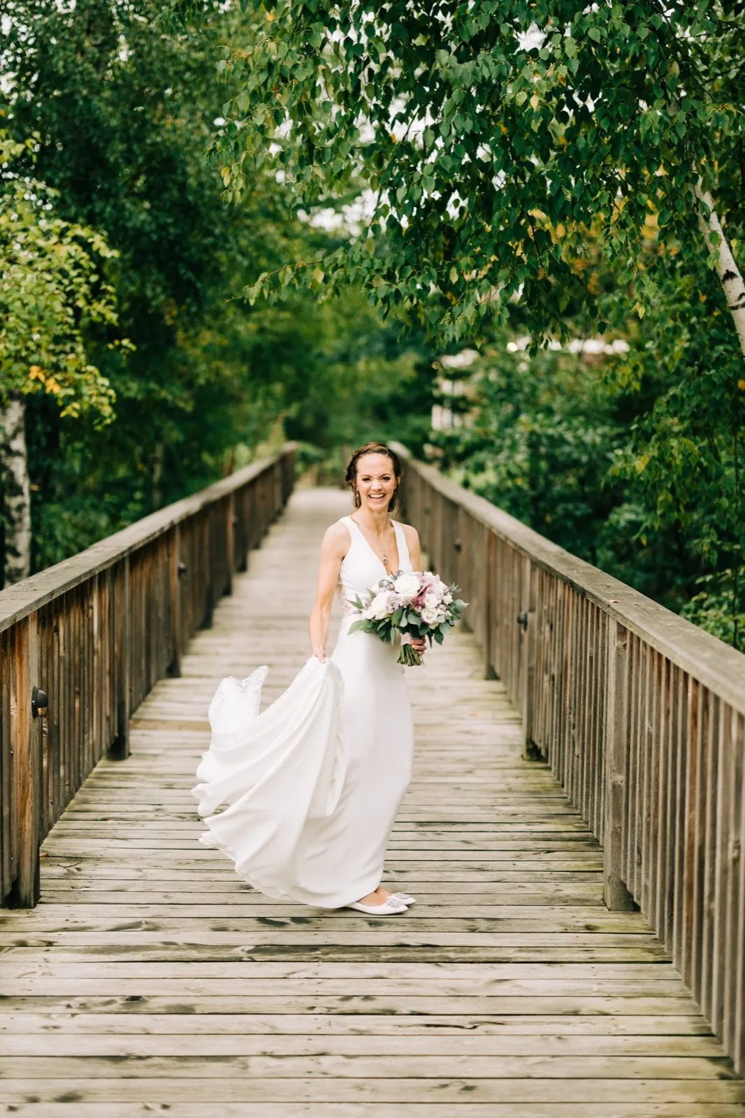 Katie laughs sweeping her dress aside on a wooden footbridge framed by birch leaves at Madden's — Tim Larsen Photography, Brainerd Lakes MN