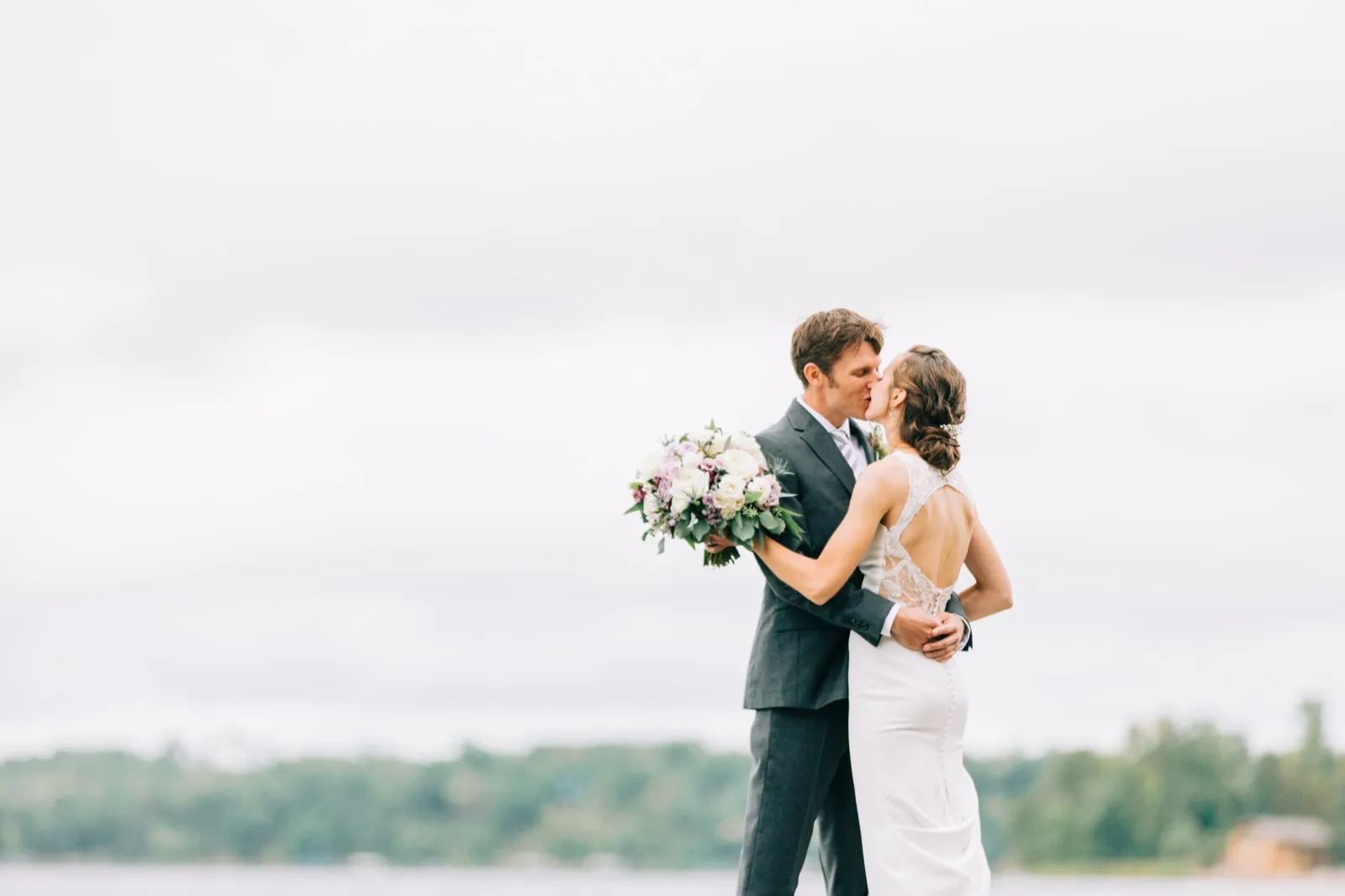 Katie and Brenden kiss on the lakeshore at Madden's, her pastel bouquet against a soft gray sky — Tim Larsen Photography, Brainerd Lakes MN