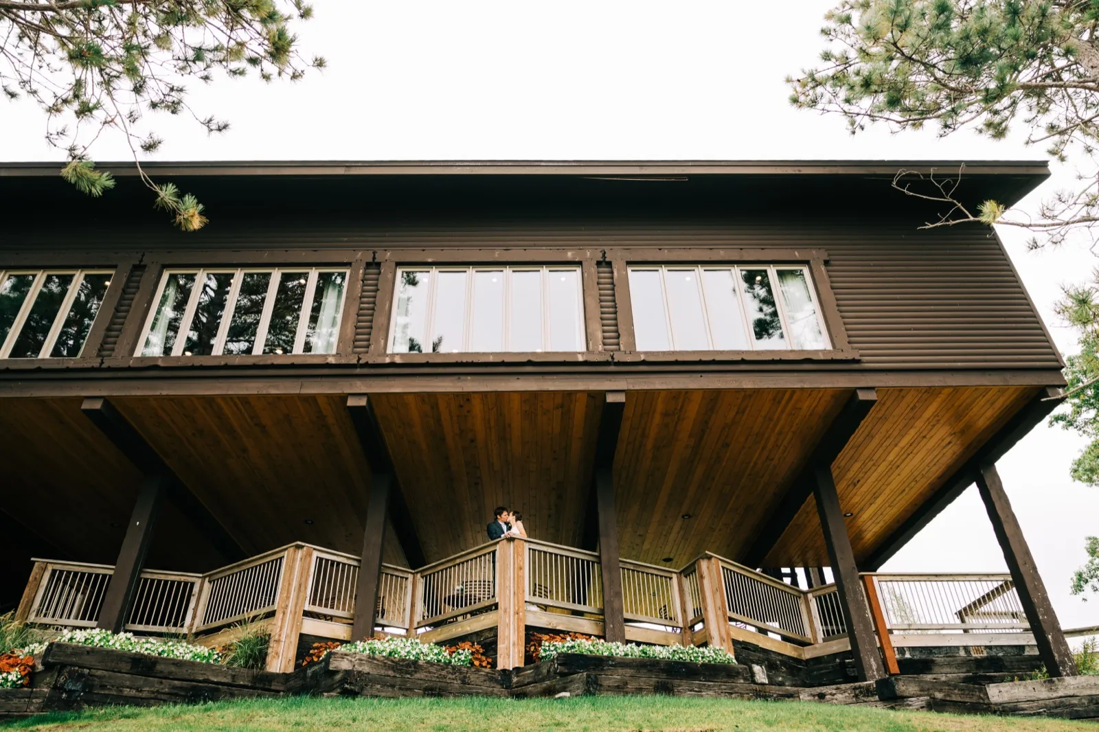 Katie and Brenden embrace on the big covered deck of a dark timber lodge at Madden's on Gull Lake, framed by pine branches — Tim Larsen Photography, Brainerd Lakes MN