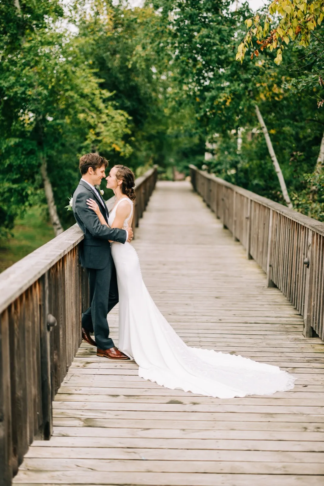 Katie and Brenden stand forehead-to-forehead on a wooden footbridge, her gown train sweeping toward the camera — Tim Larsen Photography, Brainerd Lakes MN