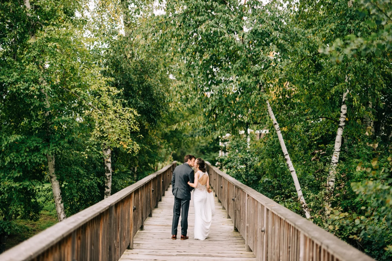 Katie and Brenden kiss at the center of a long wooden footbridge framed by birch canopy at Madden's — Tim Larsen Photography, Brainerd Lakes MN
