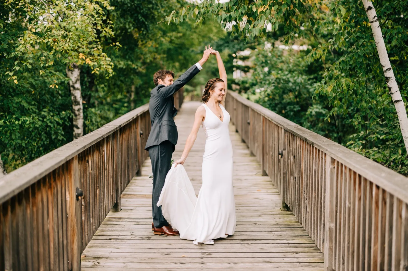 Brenden twirls Katie on a long wooden footbridge framed by birch trees, her gown trailing — Tim Larsen Photography, Brainerd Lakes MN