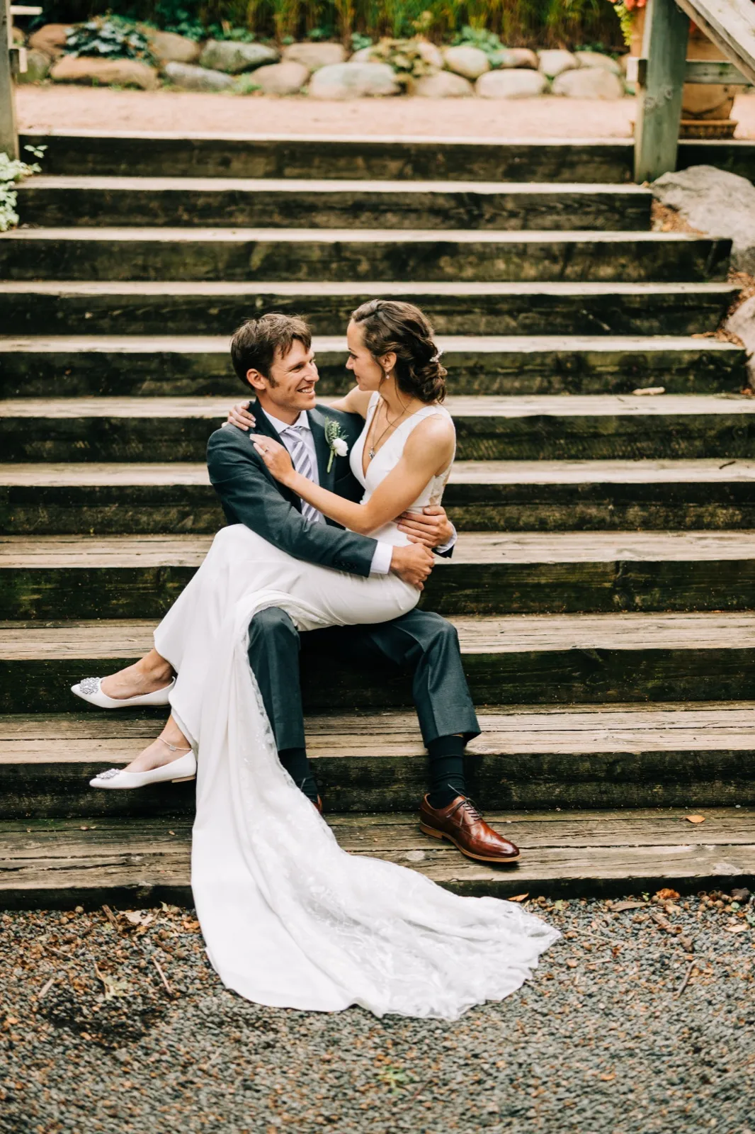 Katie sits on Brenden's lap on a wide flight of timber steps, her train spilling down the stairs as they smile at each other — Tim Larsen Photography, Brainerd Lakes MN