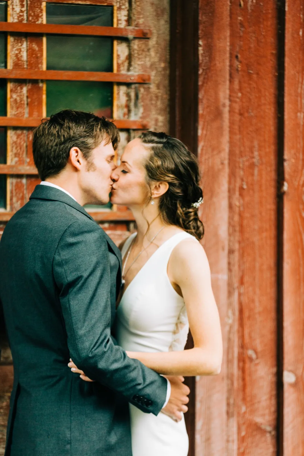 Katie and Brenden kiss against a weathered red wooden wall with a paned window at Madden's on Gull Lake — Tim Larsen Photography, Brainerd Lakes MN