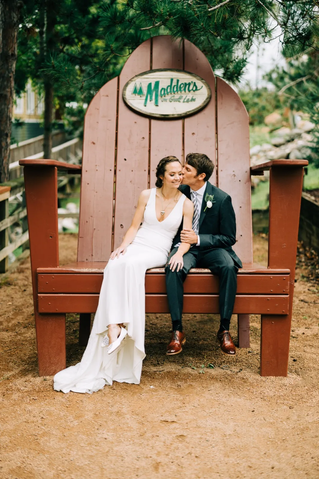 Katie and Brenden sit together in a giant red Adirondack chair bearing the Madden's on Gull Lake sign as Brenden leans in to kiss her cheek — Tim Larsen Photography, Brainerd Lakes MN
