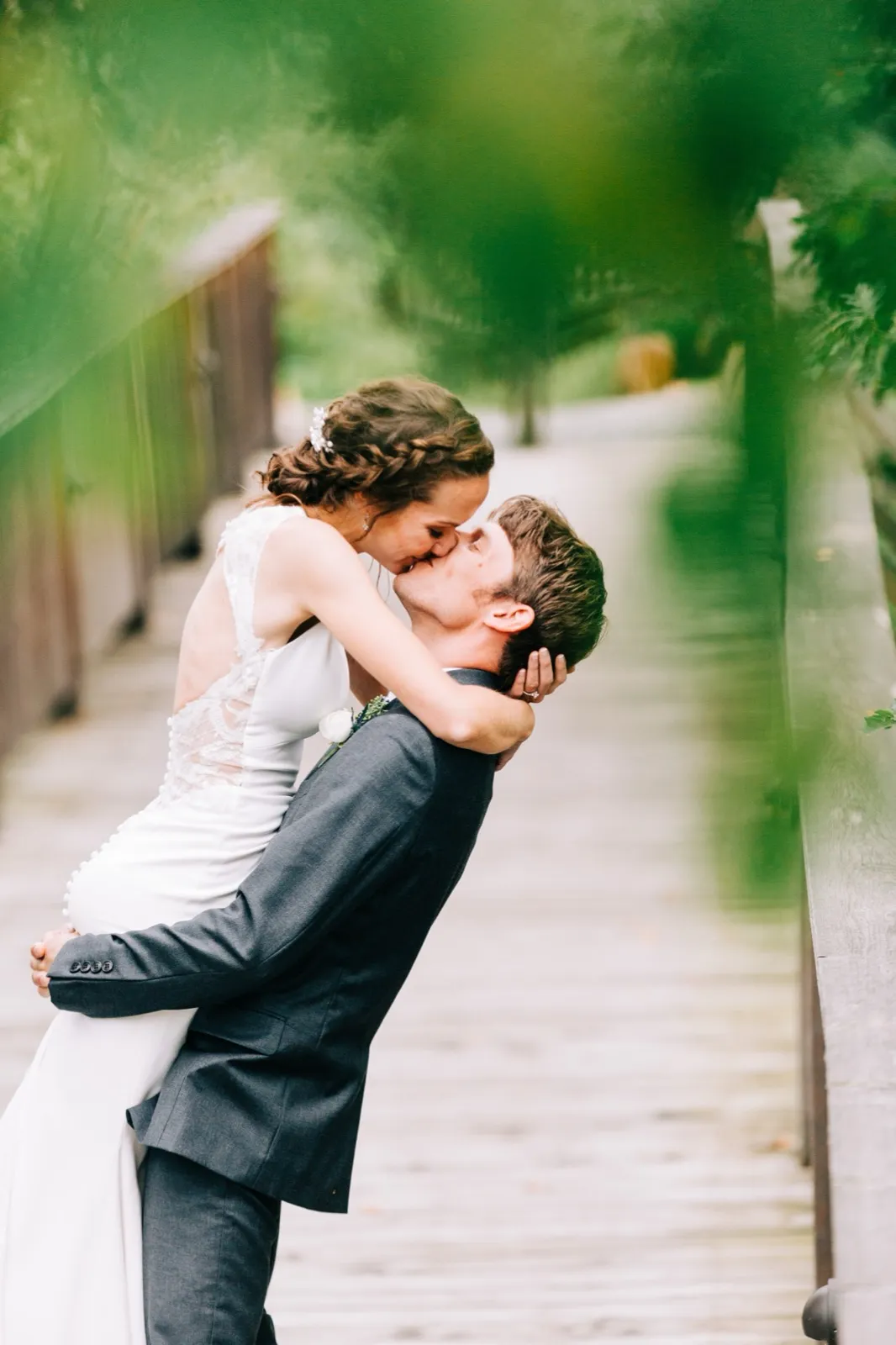 Brenden lifts Katie off the ground for a kiss on a wooden boardwalk framed by soft green foliage at Madden's — Tim Larsen Photography, Brainerd Lakes MN