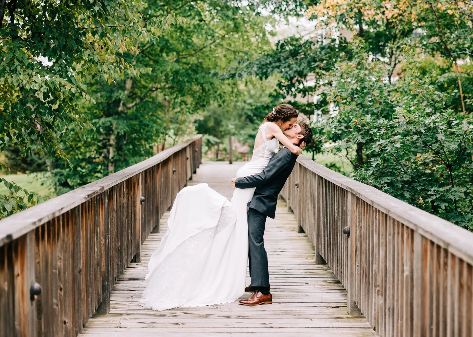 Brenden lifts Katie into a kiss at the center of a wooden footbridge at Madden's, her gown swinging across the planks — Tim Larsen Photography, Brainerd Lakes MN