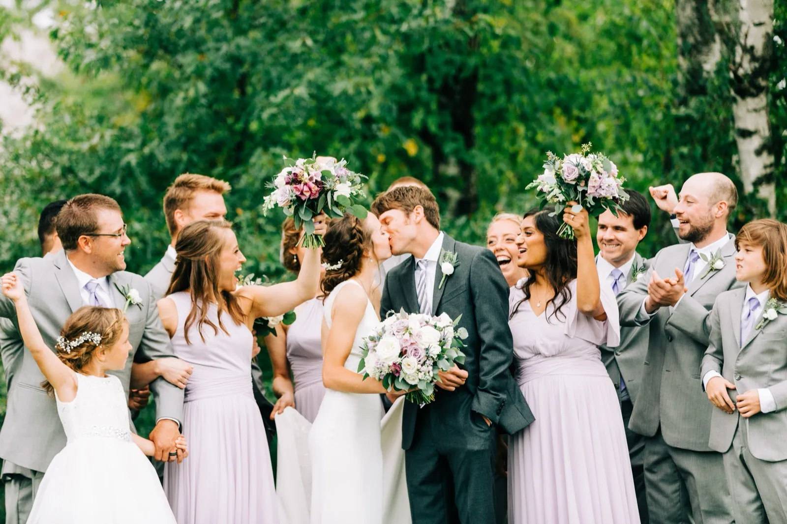 Katie and Brenden kiss at the center of their bridal party, who cheer and raise lavender bouquets on a birch-lined lawn — Tim Larsen Photography, Brainerd Lakes MN