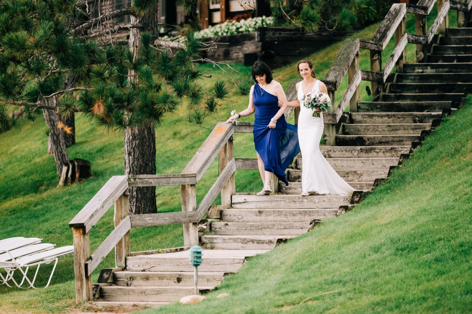 Katie walks down the long wooden hillside staircase toward the ceremony arm in arm with her mom in a royal-blue dress — Tim Larsen Photography, Brainerd Lakes MN