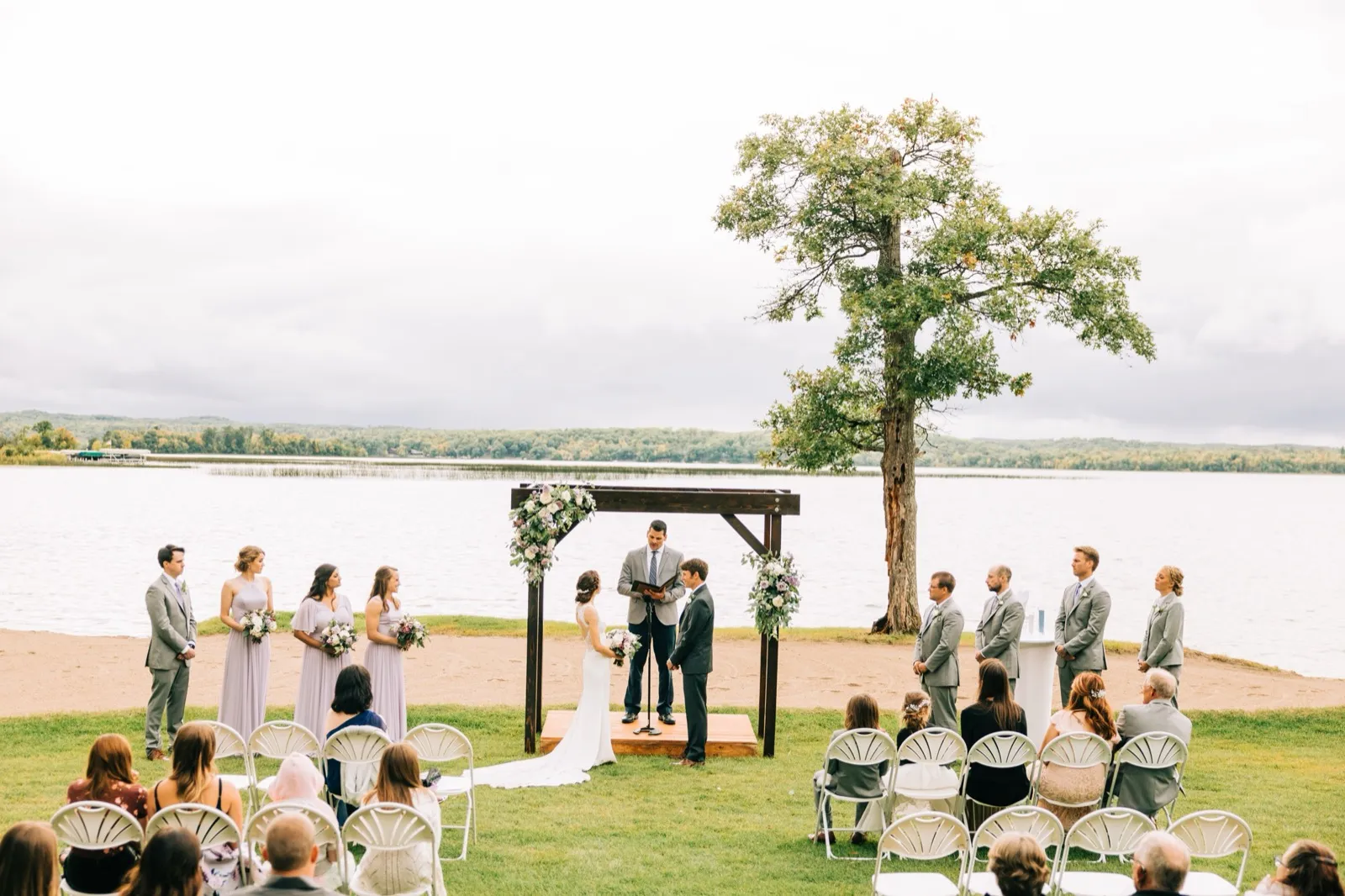 Katie and Brenden's ceremony beneath a floral wooden arbor on the lawn beside Gull Lake, the bridal party flanking them under a large oak — Tim Larsen Photography, Brainerd Lakes MN