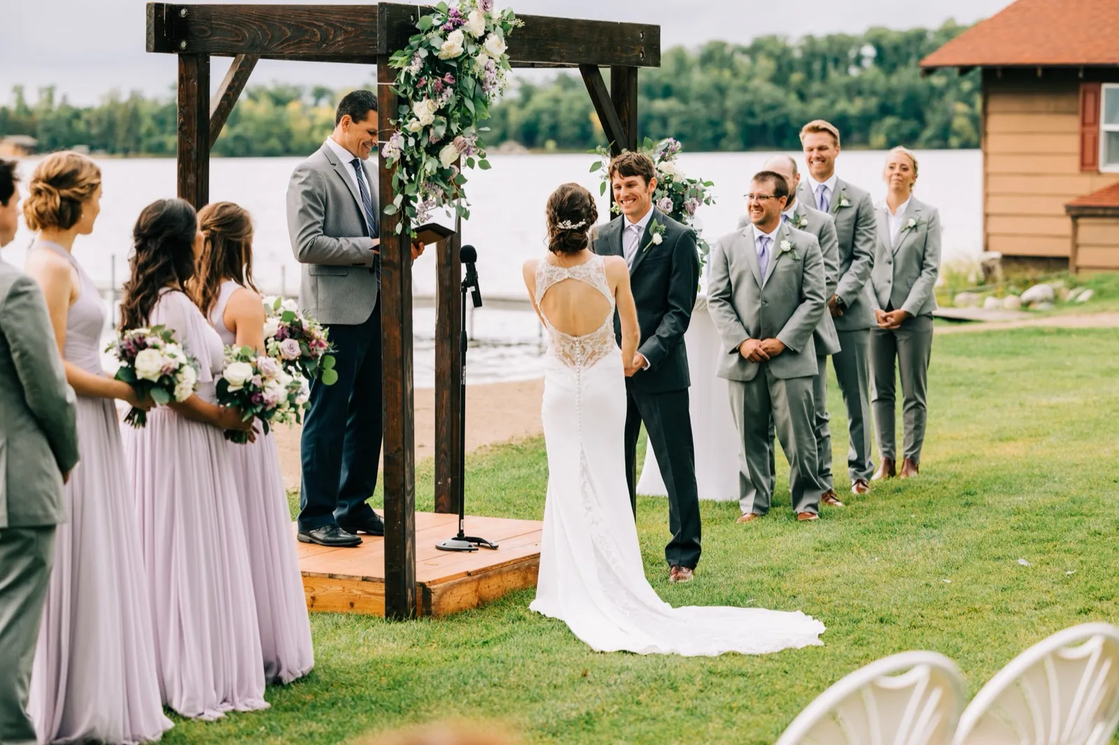 Katie and Brenden hold hands beneath a wooden arch draped with lavender and white florals, Gull Lake and a boathouse behind them — Tim Larsen Photography, Brainerd Lakes MN