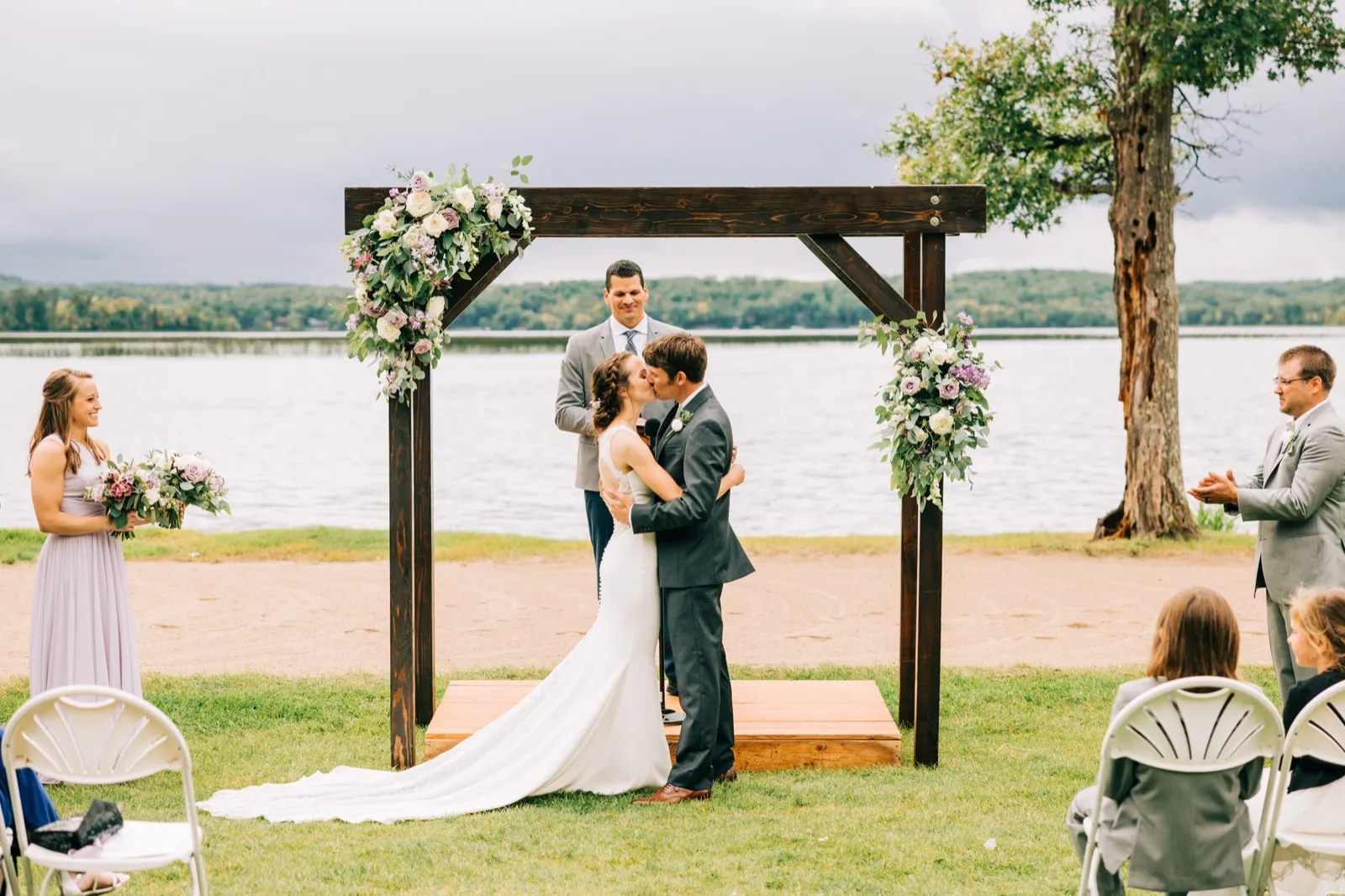 Katie and Brenden share their first kiss beneath a wooden arch draped with lavender and cream florals, Gull Lake behind them — Tim Larsen Photography, Brainerd Lakes MN