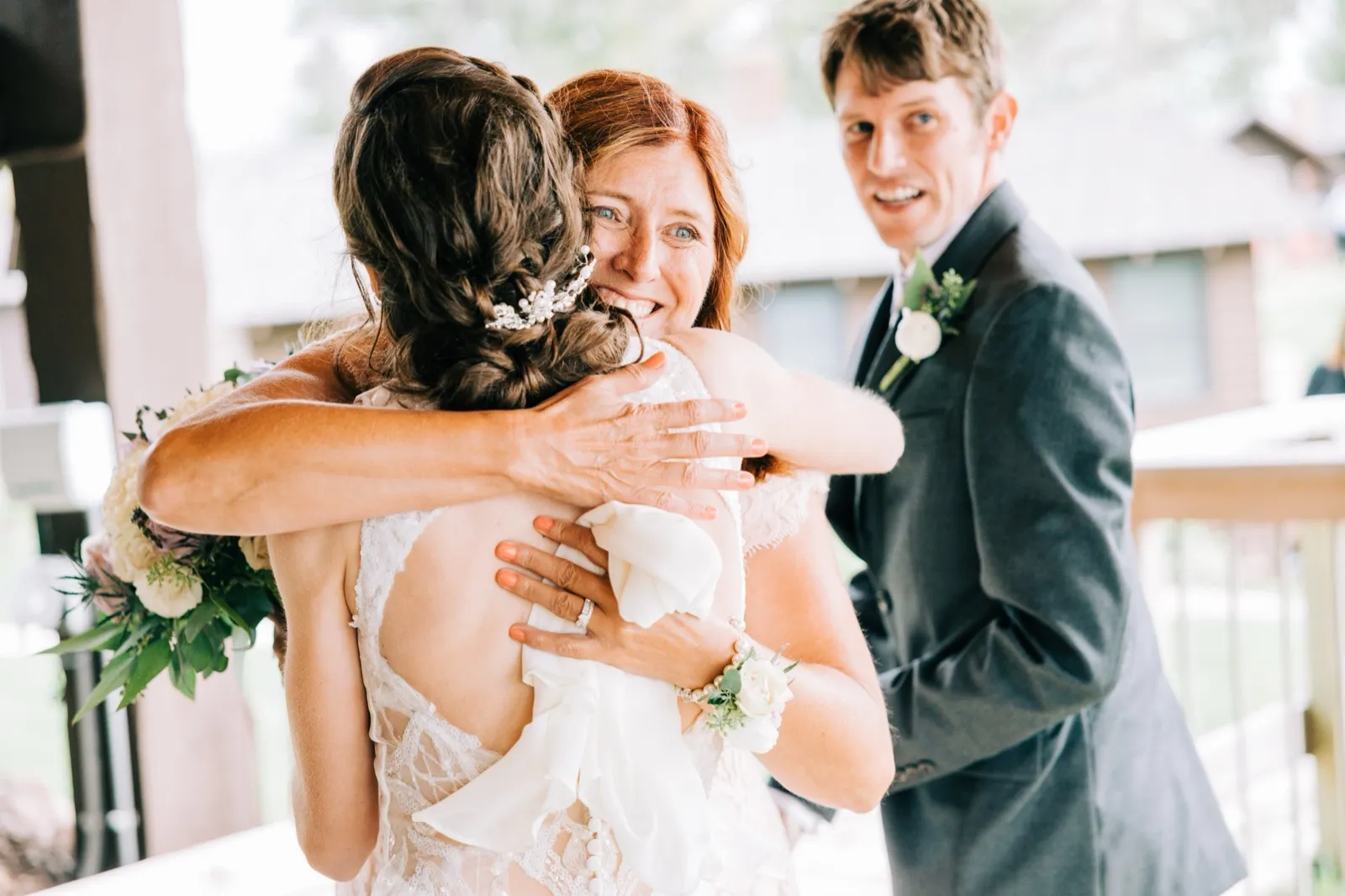 A woman embraces Katie from behind while Brenden stands smiling nearby on a covered porch at Madden's — Tim Larsen Photography, Brainerd Lakes MN