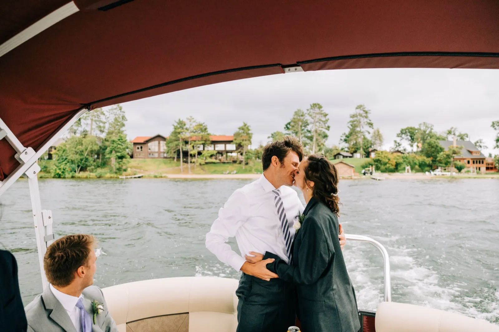 Katie, in Brenden's suit jacket, kisses Brenden under the burgundy canopy of a pontoon on Gull Lake as a groomsman sits in the foreground — Tim Larsen Photography, Brainerd Lakes MN