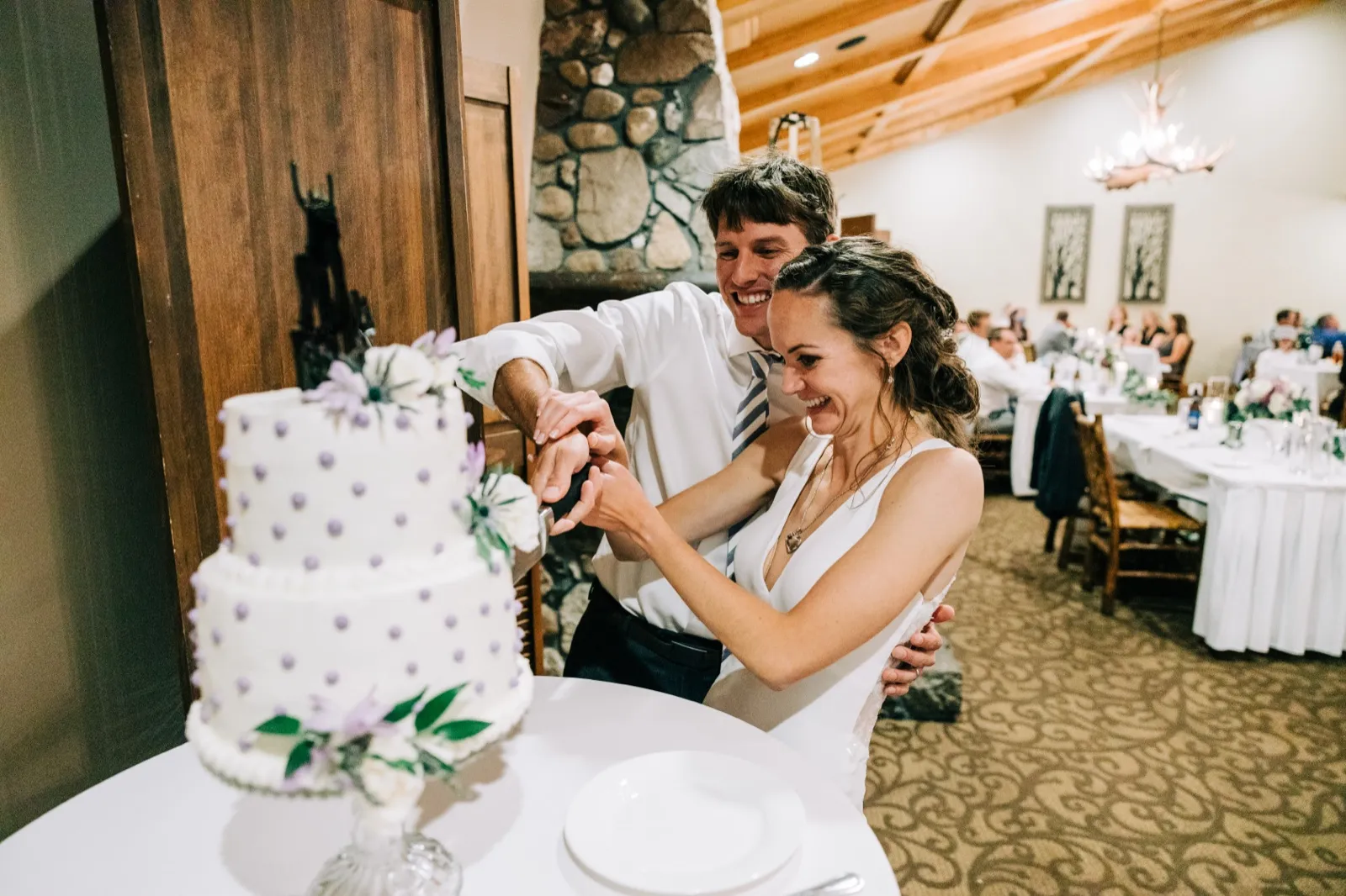 Katie and Brenden laugh as they cut their lavender-dotted wedding cake in front of a stone fireplace — Tim Larsen Photography, Brainerd Lakes MN