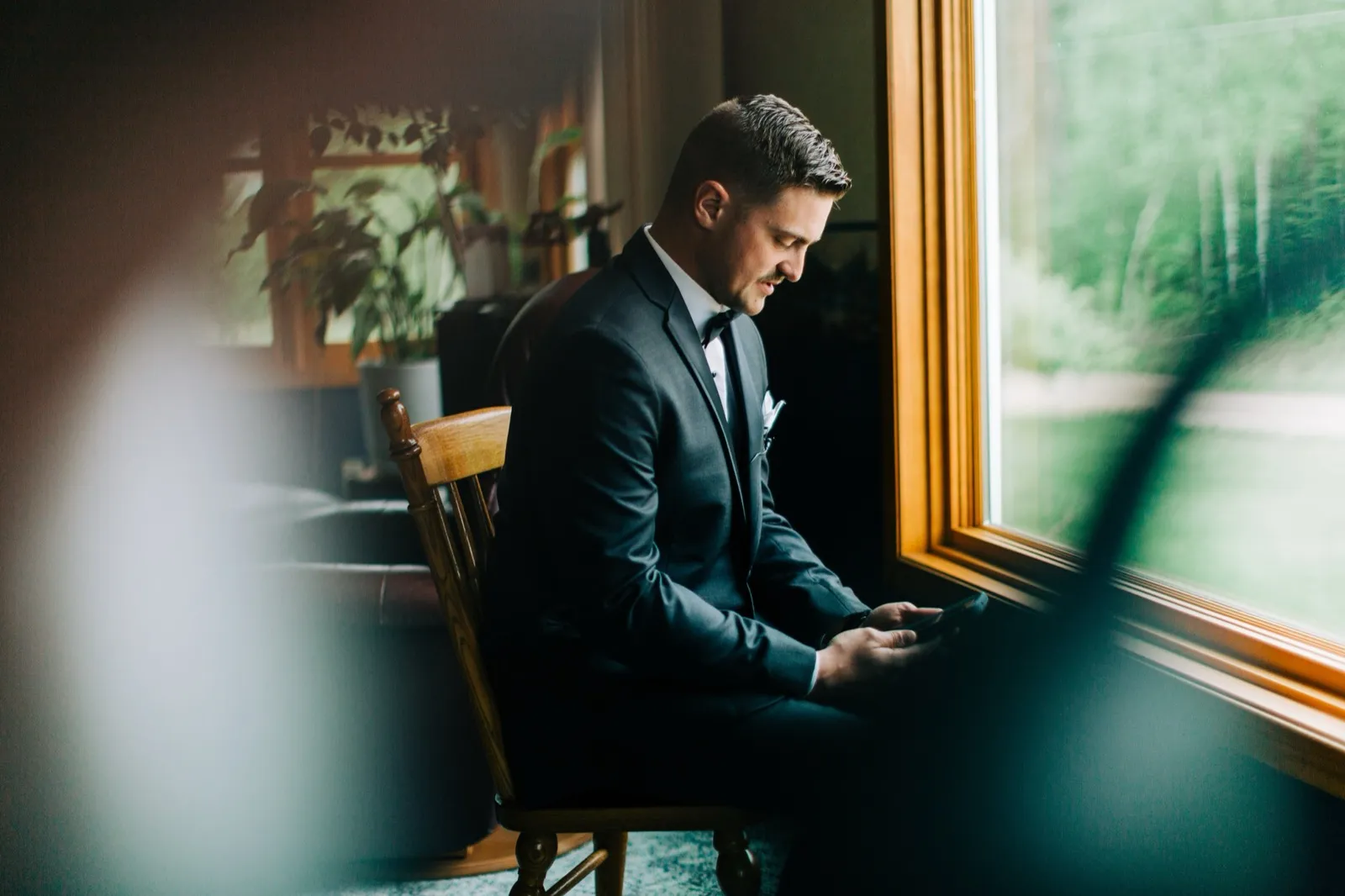 Jonathon sits by a large window in his suit, reading a few last messages in soft side light — Tim Larsen Photography, Brainerd Lakes MN