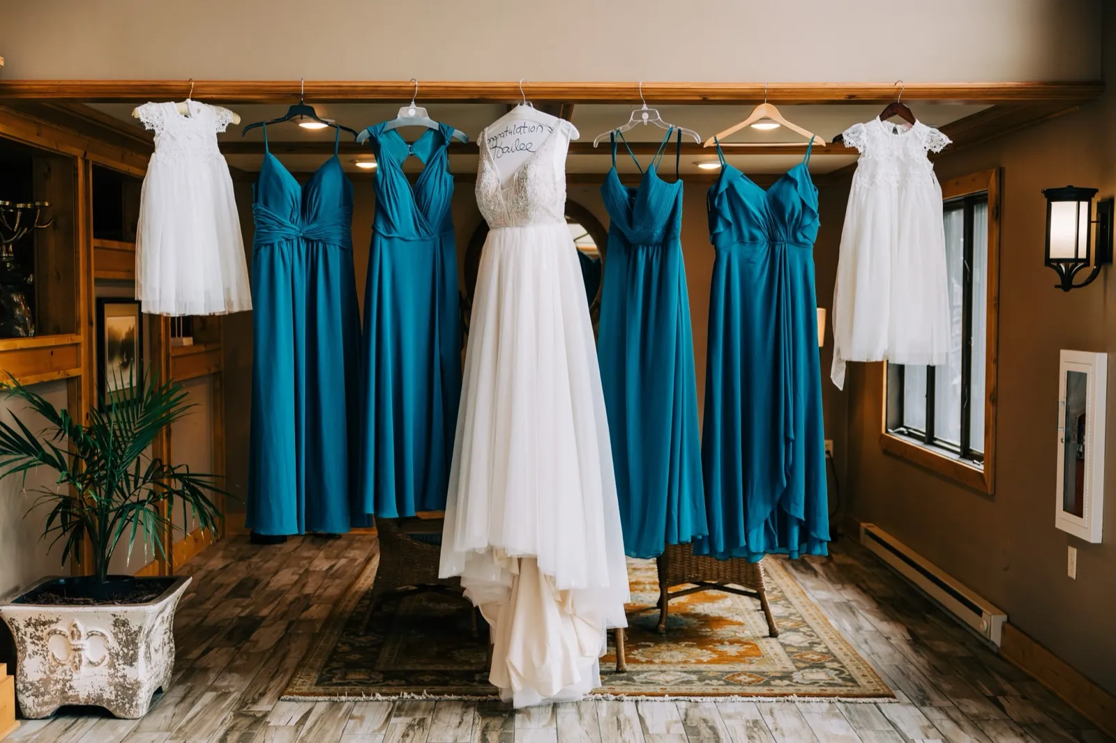 Hailee's gown hanging between five teal bridesmaid dresses and two flower-girl dresses on a wooden beam at Madden's — Tim Larsen Photography, Brainerd Lakes MN
