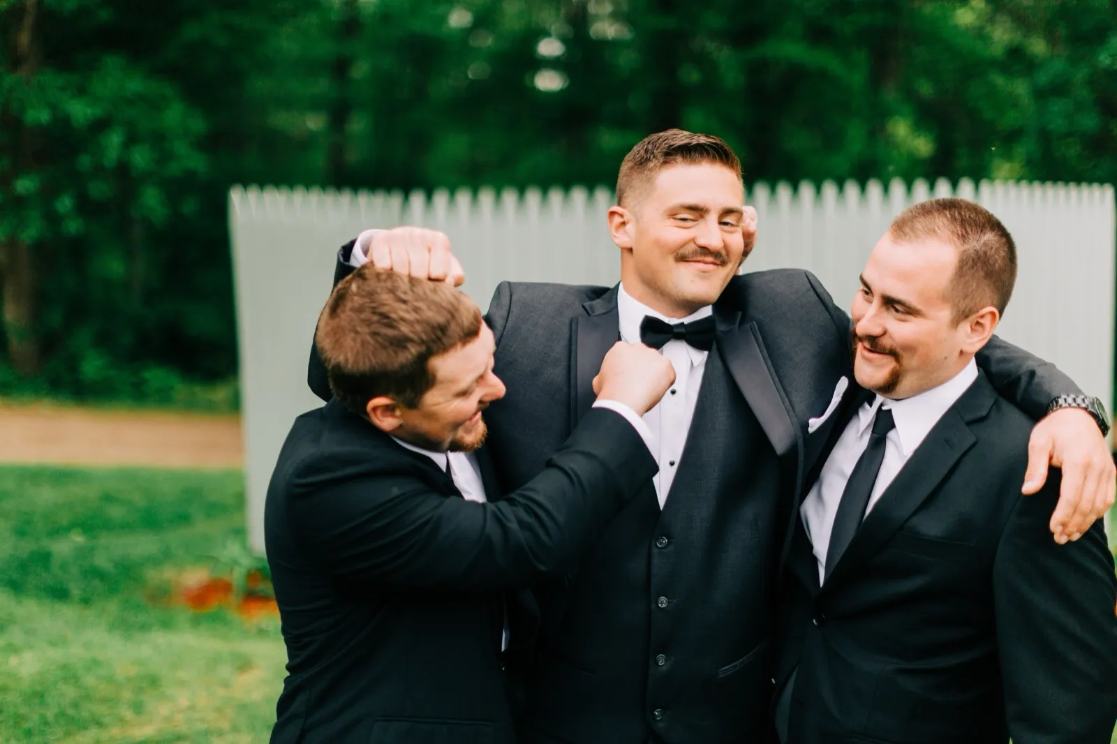 Jonathon smirks as two groomsmen flank him with arms over his shoulders in front of a white fence and the forest — Tim Larsen Photography, Brainerd Lakes MN