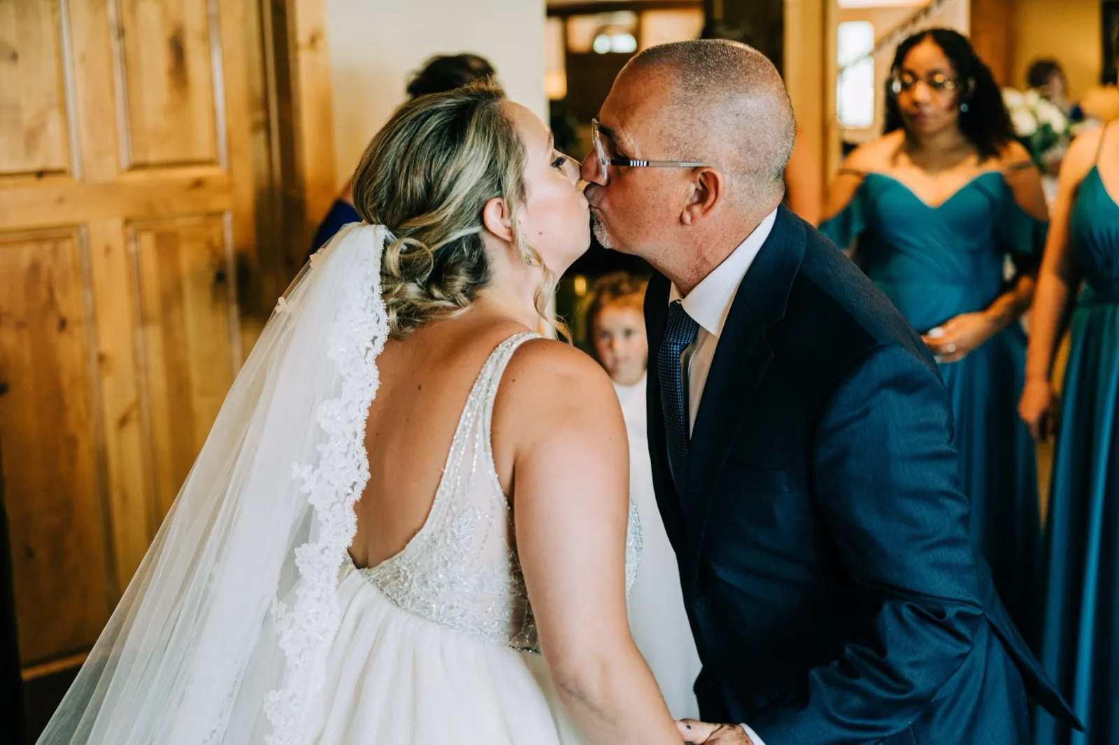 Hailee and her father share a kiss as a small flower girl peeks between them before the ceremony — Tim Larsen Photography, Brainerd Lakes MN