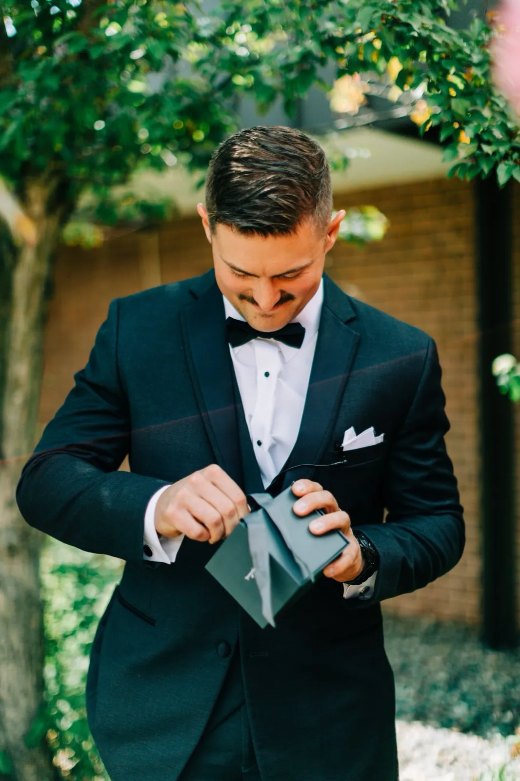 Jonathon smiles as he opens a small gift box beneath green tree foliage before the ceremony — Tim Larsen Photography, Brainerd Lakes MN