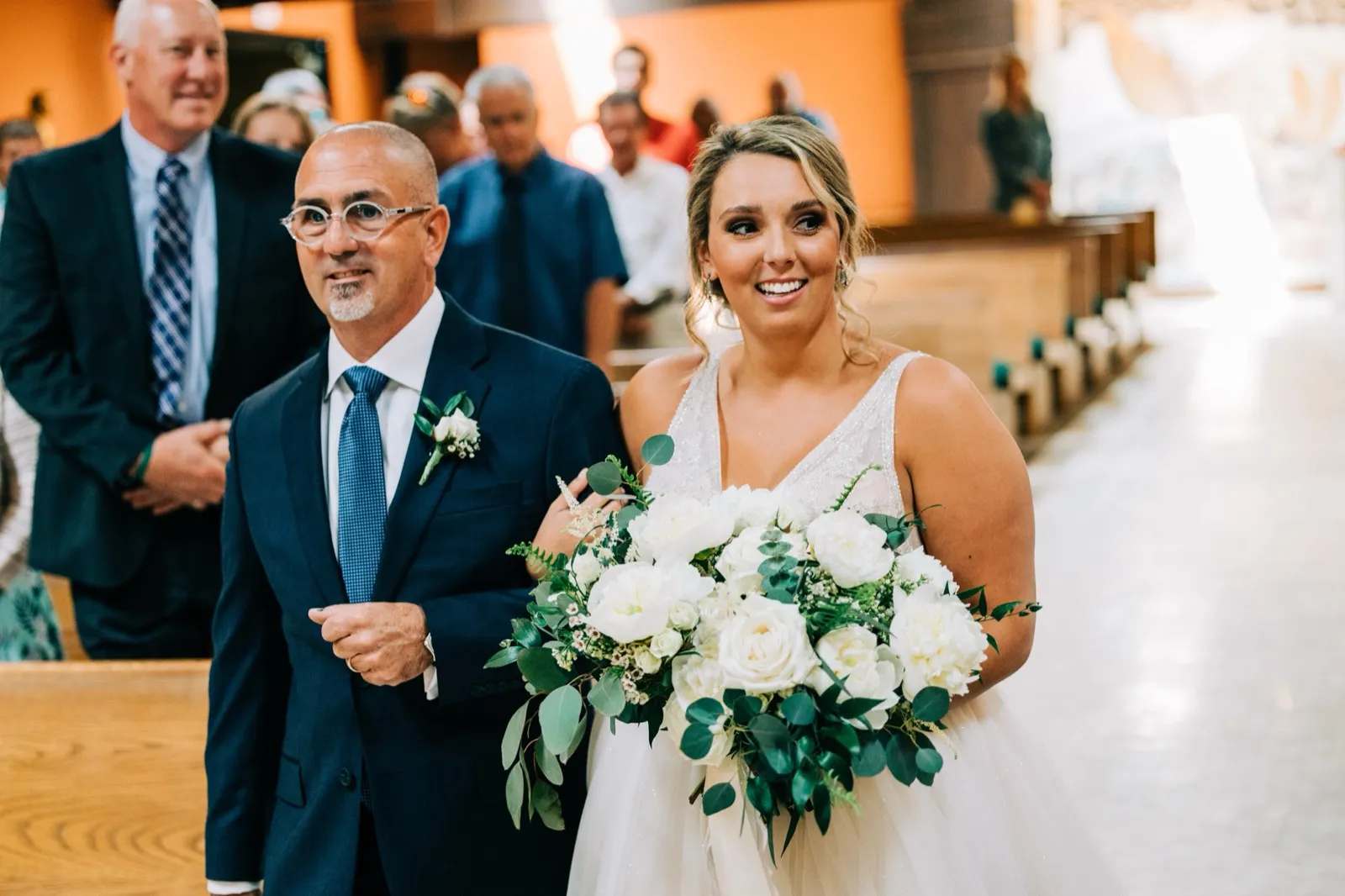 Hailee walks down the church aisle on her father's arm, beaming, a smiling guest just behind — Tim Larsen Photography, Brainerd Lakes MN