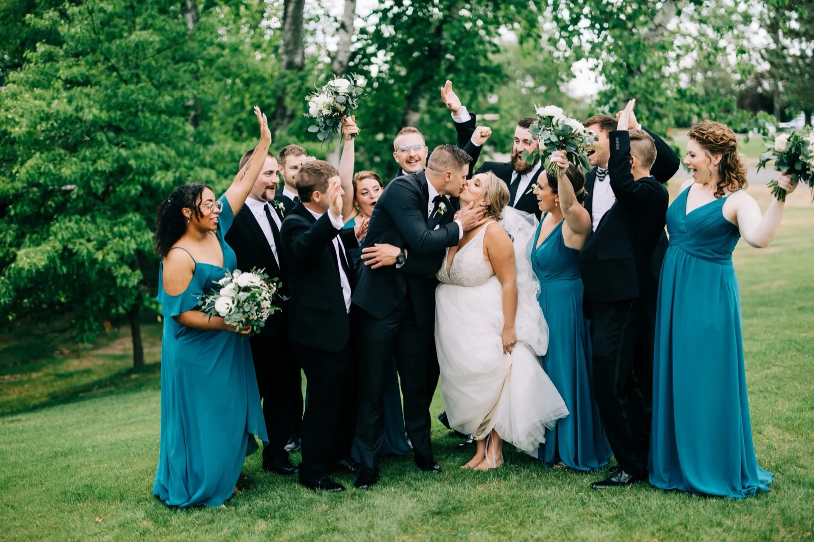 Hailee and Jonathon kiss at the center of their bridal party, who throw their arms up cheering on a green lawn at Madden's — Tim Larsen Photography, Brainerd Lakes MN