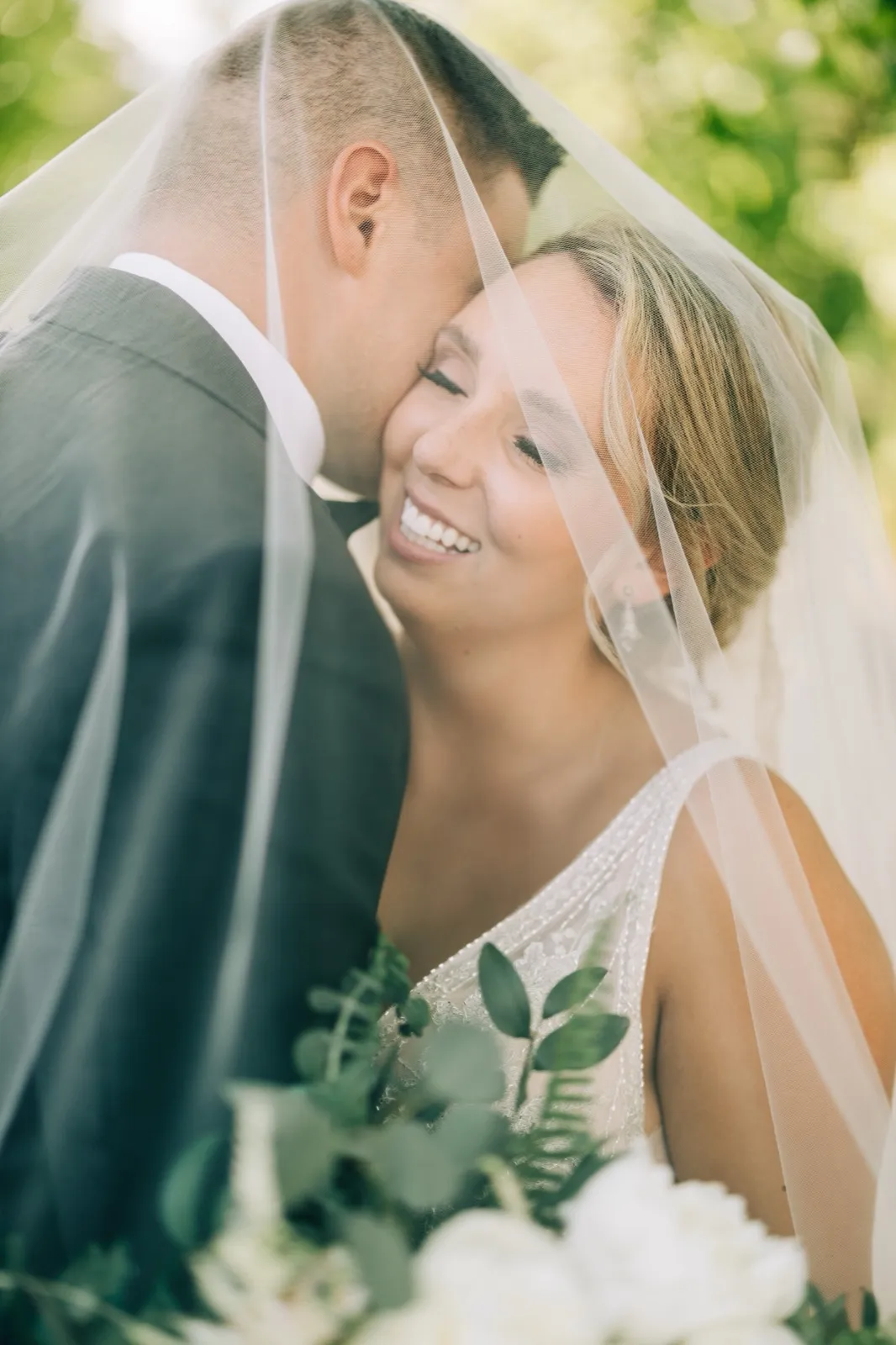 Hailee smiles under her veil as it drapes around her and Jonathon, white blooms and greenery in the foreground — Tim Larsen Photography, Brainerd Lakes MN